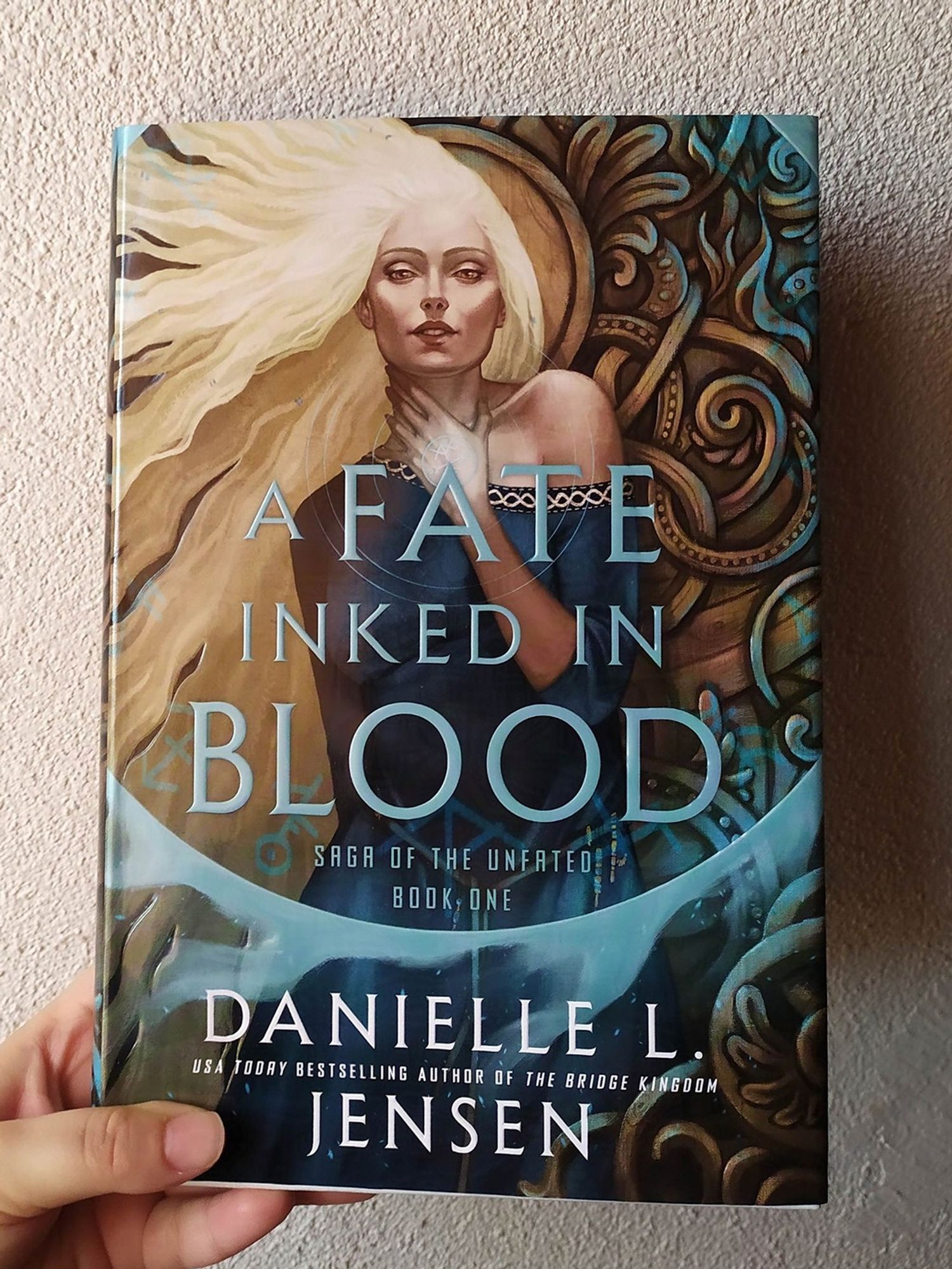 Photo of the foiled book cover of A FATE INKED IN BLOOD by Danielle L. Jensen, featuring a long-haired blonde woman with her hand over her neck. A blue circle frames her face and illuminates the norse-inspired woodwork on the background. Illustration by Eleonor Piteira, design by Ella Laytham.