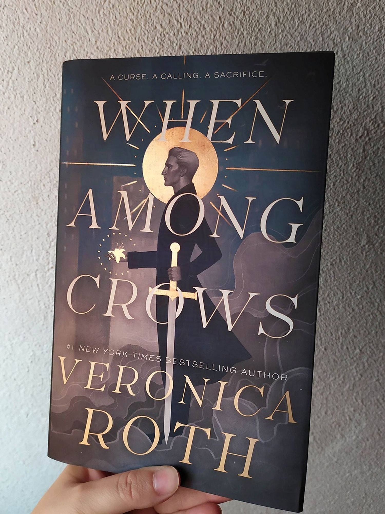 Photo of the book cover of WHEN AMONG CROWS by Veronica Roth, featuring a man with a gold halo holding a sword and a flower. Illustration by Eleonor Piteira, design by Katie Klimowicz.