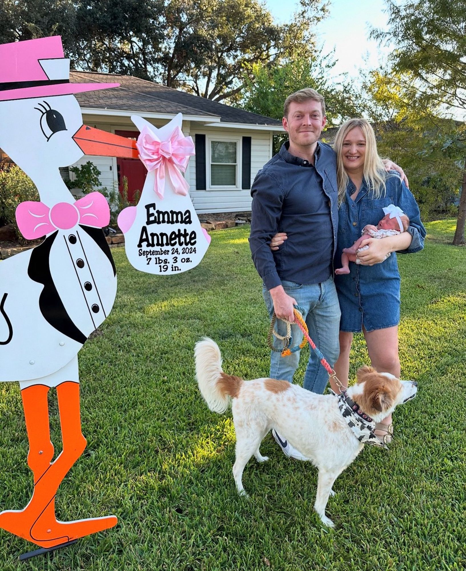 A white man & woman in a front yard holding their newborn baby. A leashed tan & white dog is in front of them, & a large wooden stork in a pink hat & tie stands on the left side. The bundle in the stork’s beak reads “Emma Annette, September 24, 2024, 7 lbs. 3 oz., 19 in”