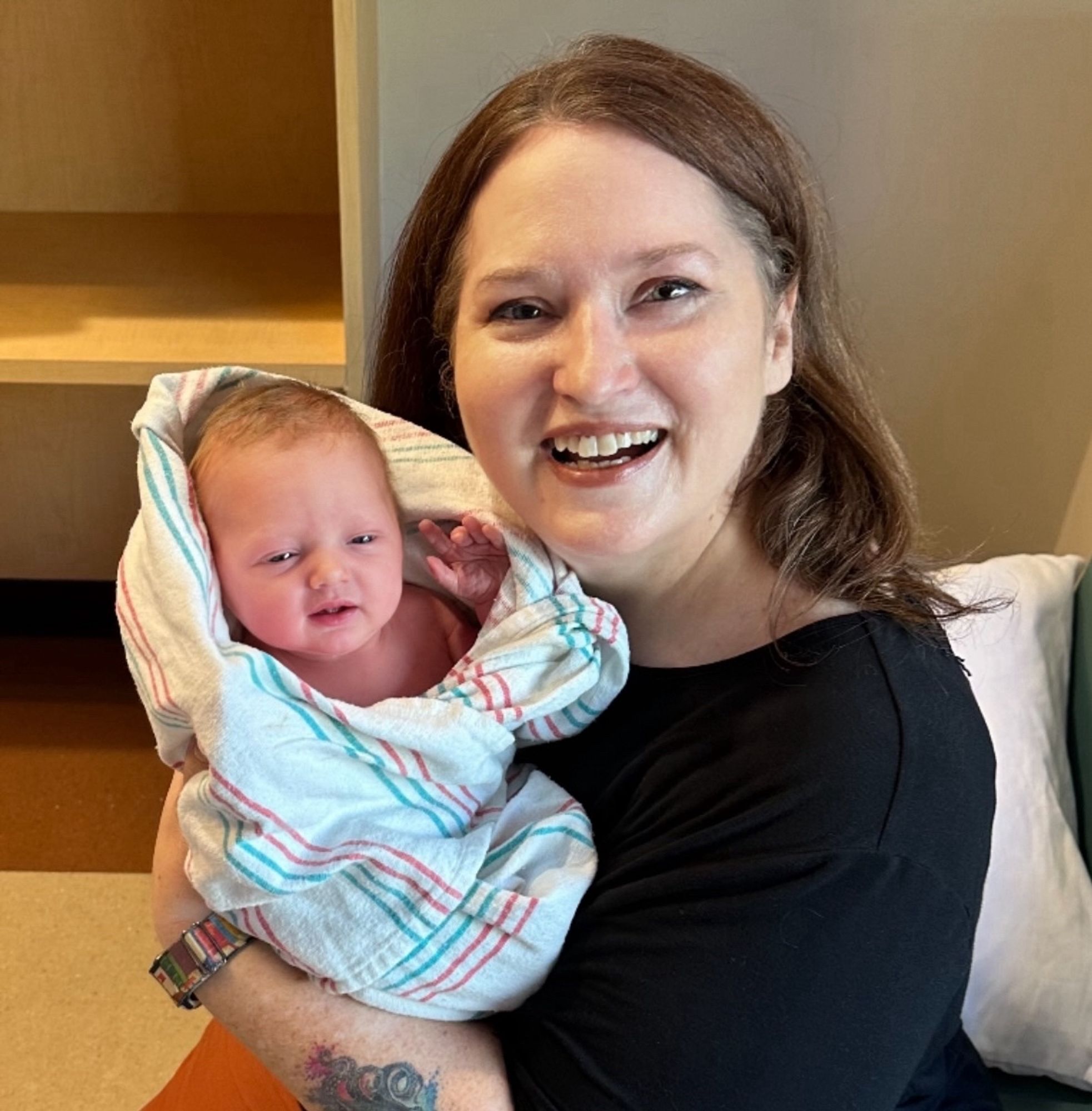 A white woman with longish brown hair wearing a black shirt is holding up a newborn baby girl wrapped in a blanket