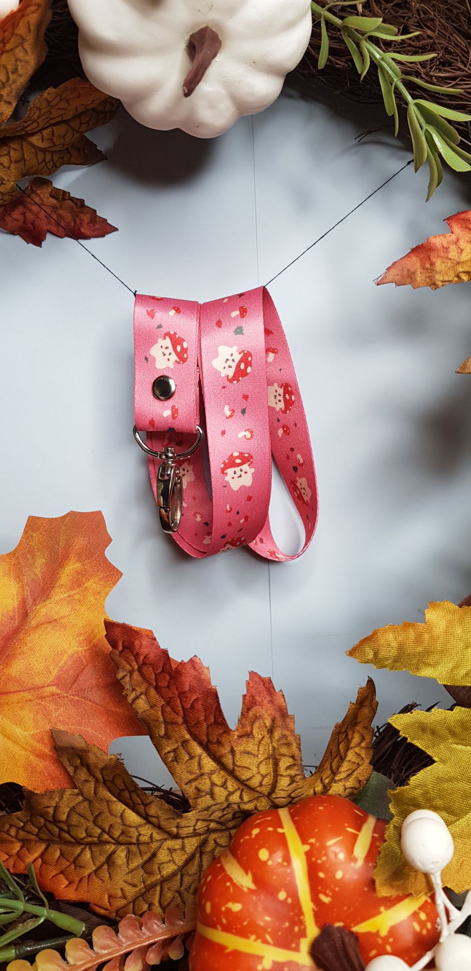 a photo of a pink laynard with red and white capped mushroom art on it. it is hanging from a string in the middle of an autumn wreath.