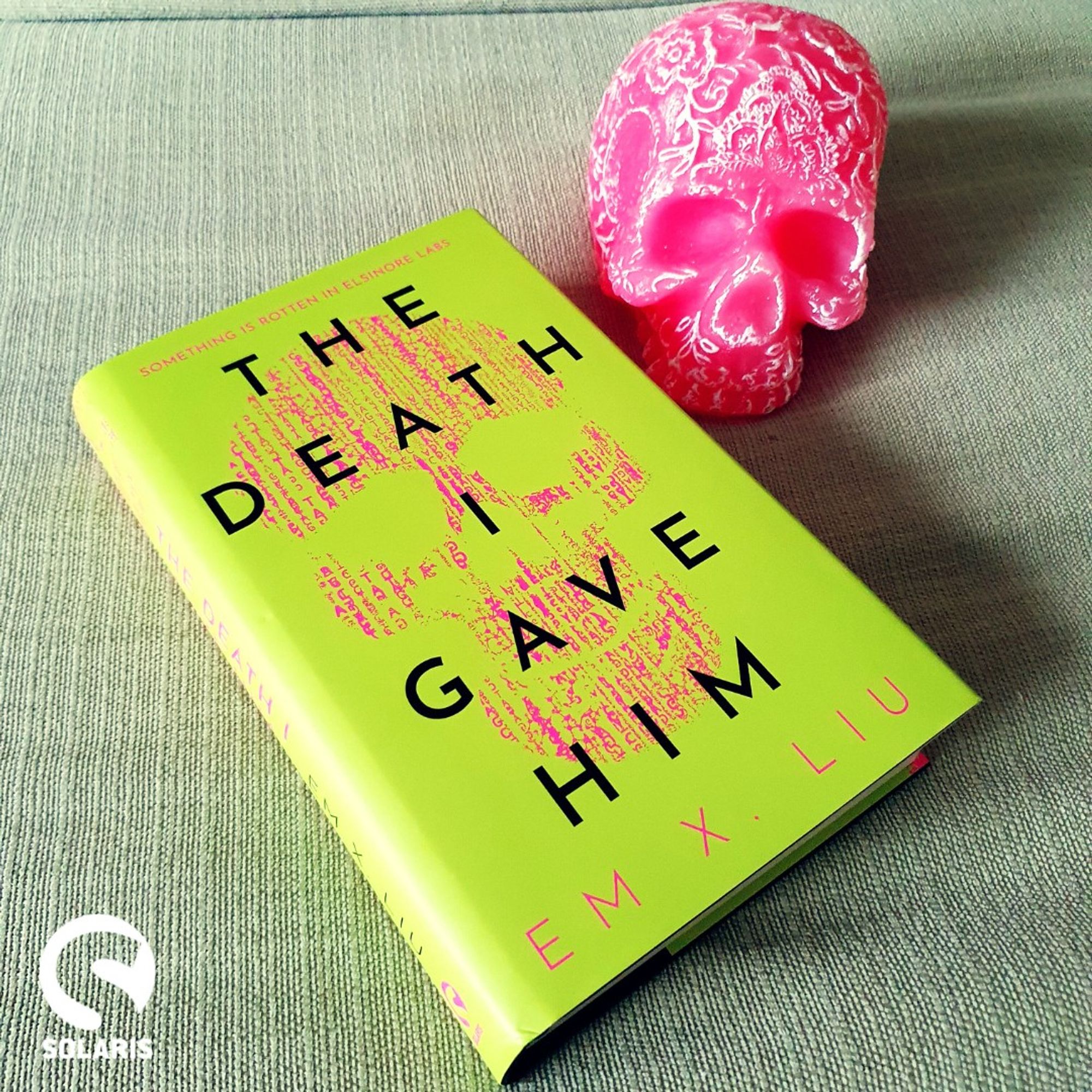 Against a pale fabric background with the white Solaris logo in the bottom left corner, lies a hardback copy of THE DEATH I GAVE HIM by Em X. Liu next to a pink skull!