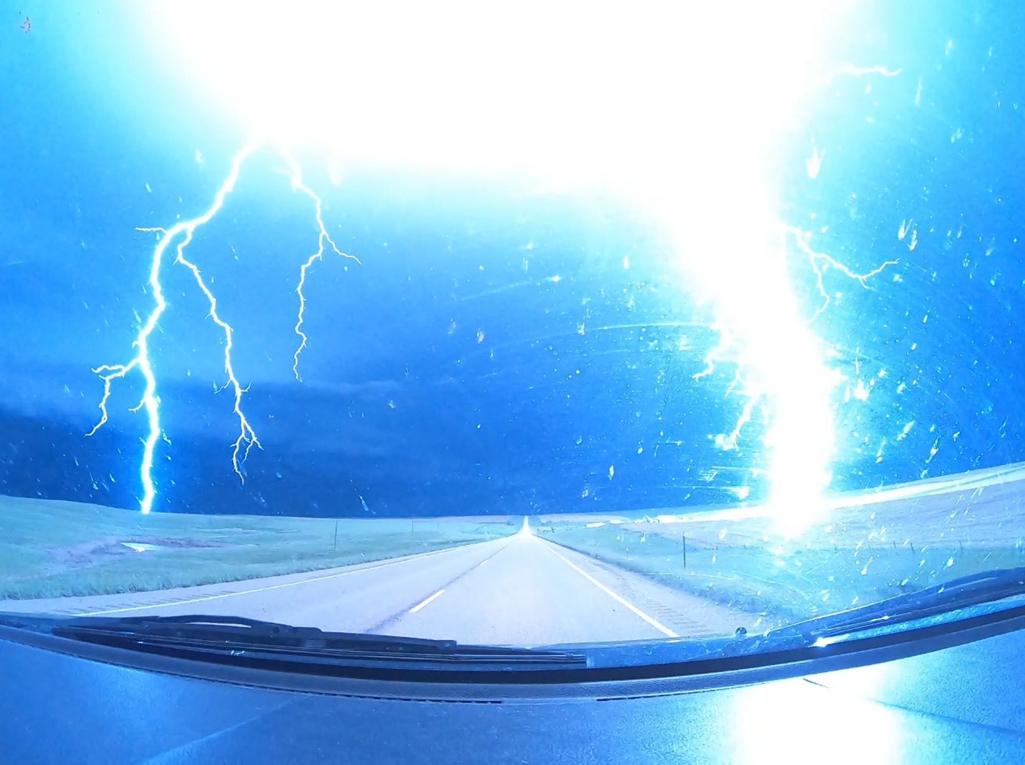 twin bolts of lightning simultaneously striking either side of the road from the point of view of a dash cam. Vibrant blue is the dominant color