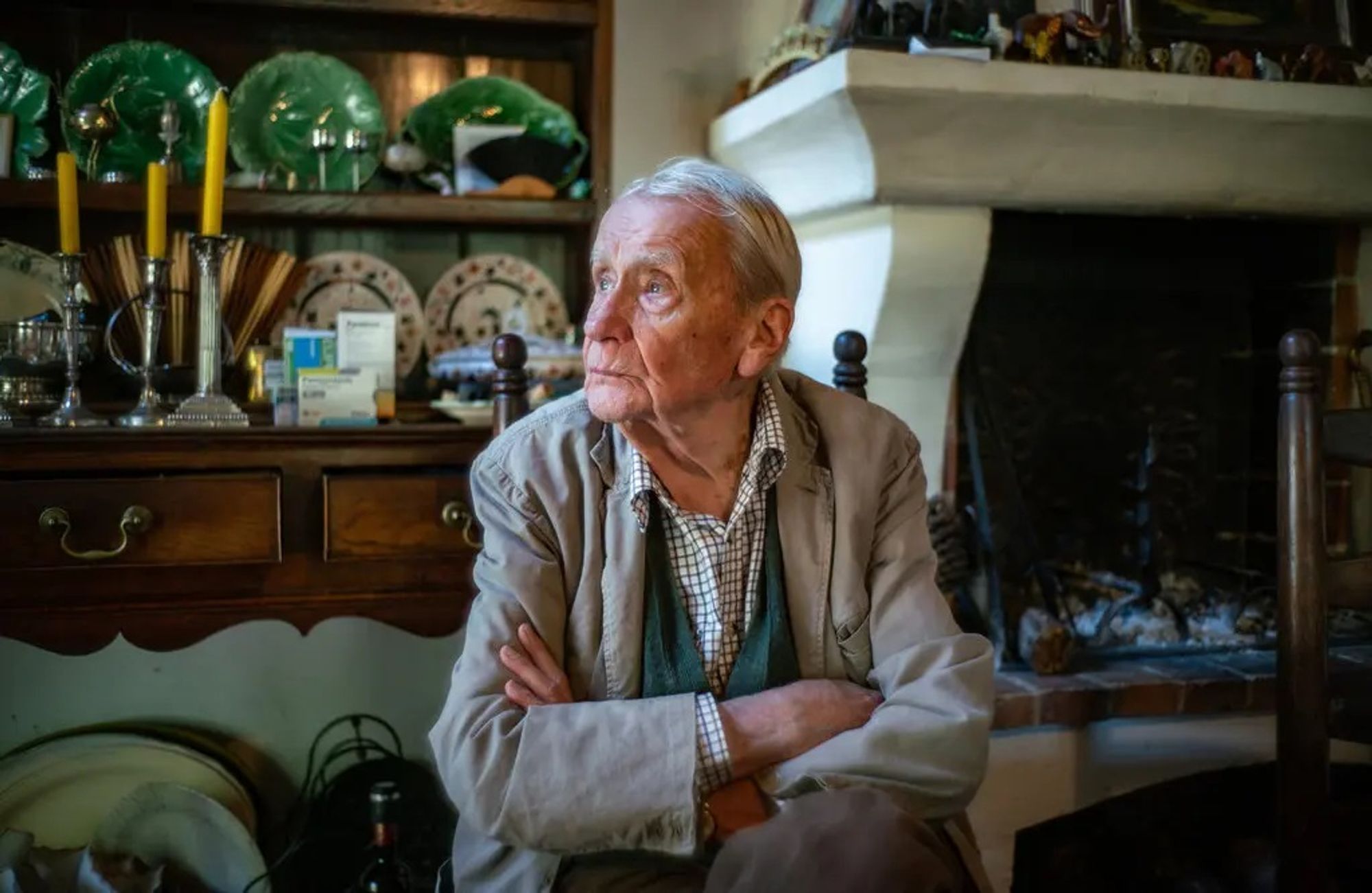 Christopher Tolkien sat in his living room, arms folded, and a large fireplace is behind him along with a dressing table with candles and plates.