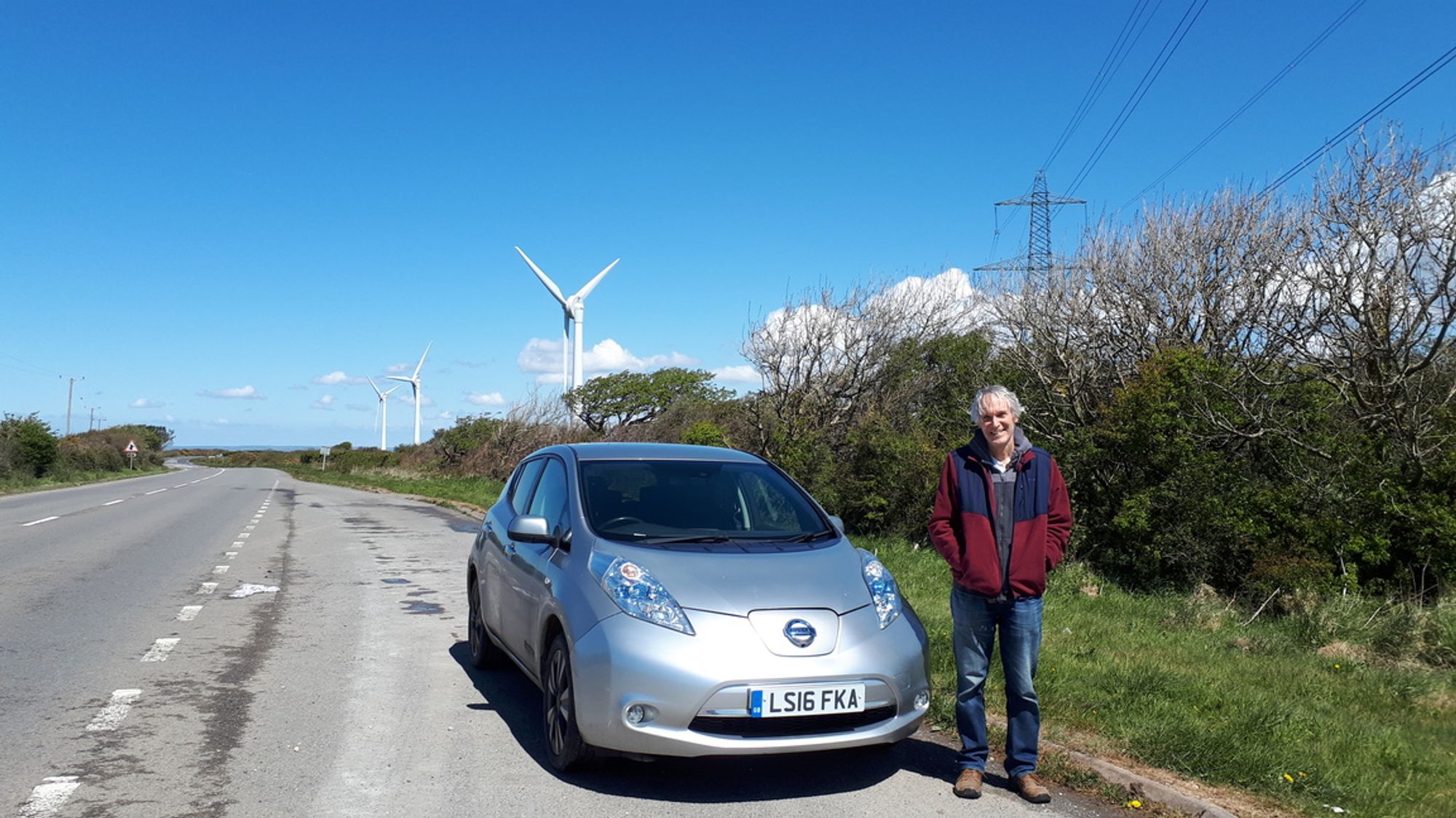 A Nissan LEAF vehicle-to-grid enabled electric vehicle, pictured in front of Otterham Down wind farm and some UK transmission grid pylons