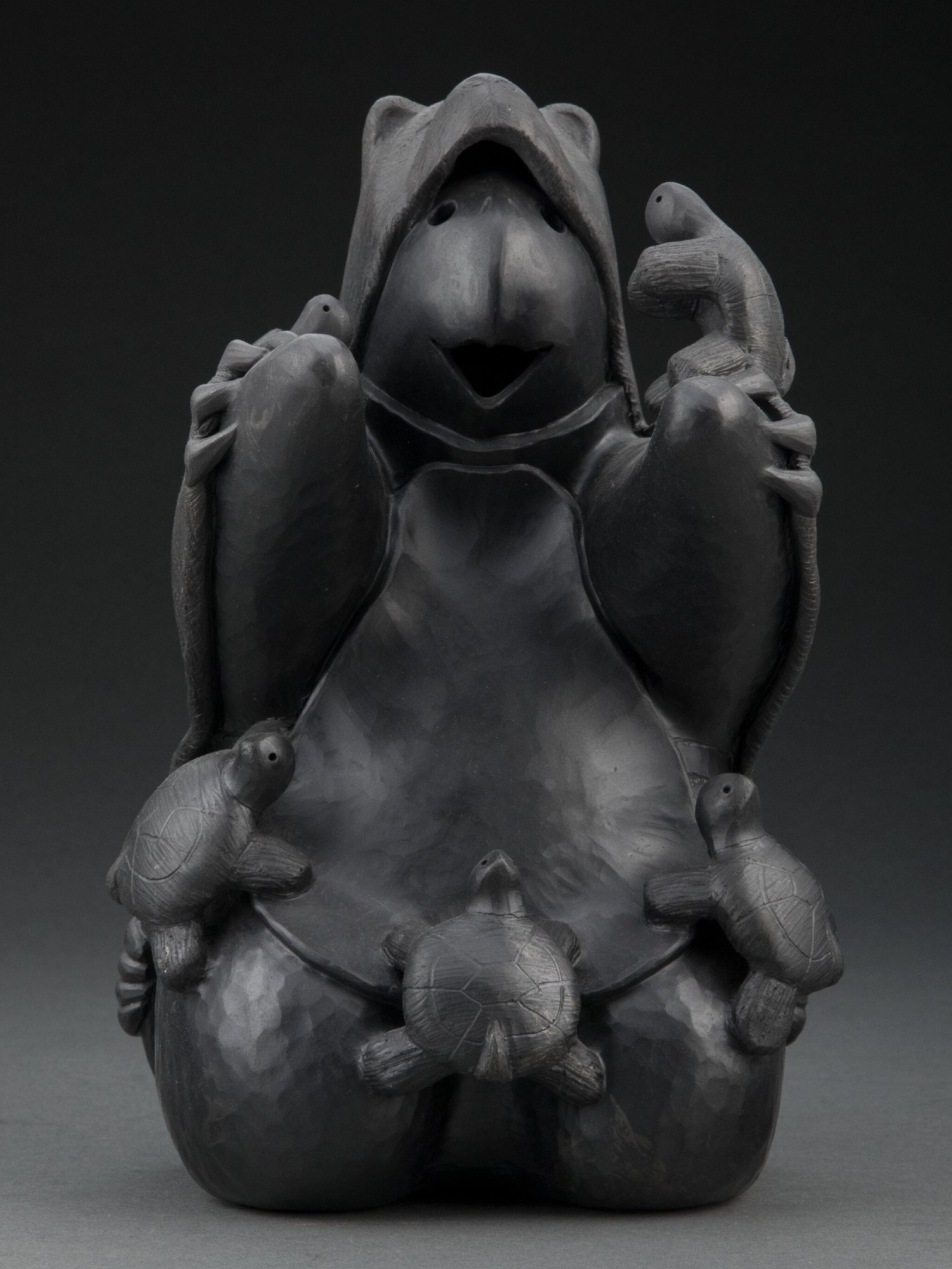 official auction photo of the object, front view on gradient grey-black background: blackware ceramic sculpture depicting a turtle storyteller (sitting upright with mouth open as if speaking) dressed in a bear hide, surrounded by small turtle children listening to it talk

"Randy's artistic journey primarily centers around the turtle, symbolizing the Choctaw's storytellers, and the bear, representing the strength and courage of the male facet of the tribe."
https://randychitto.com/bio