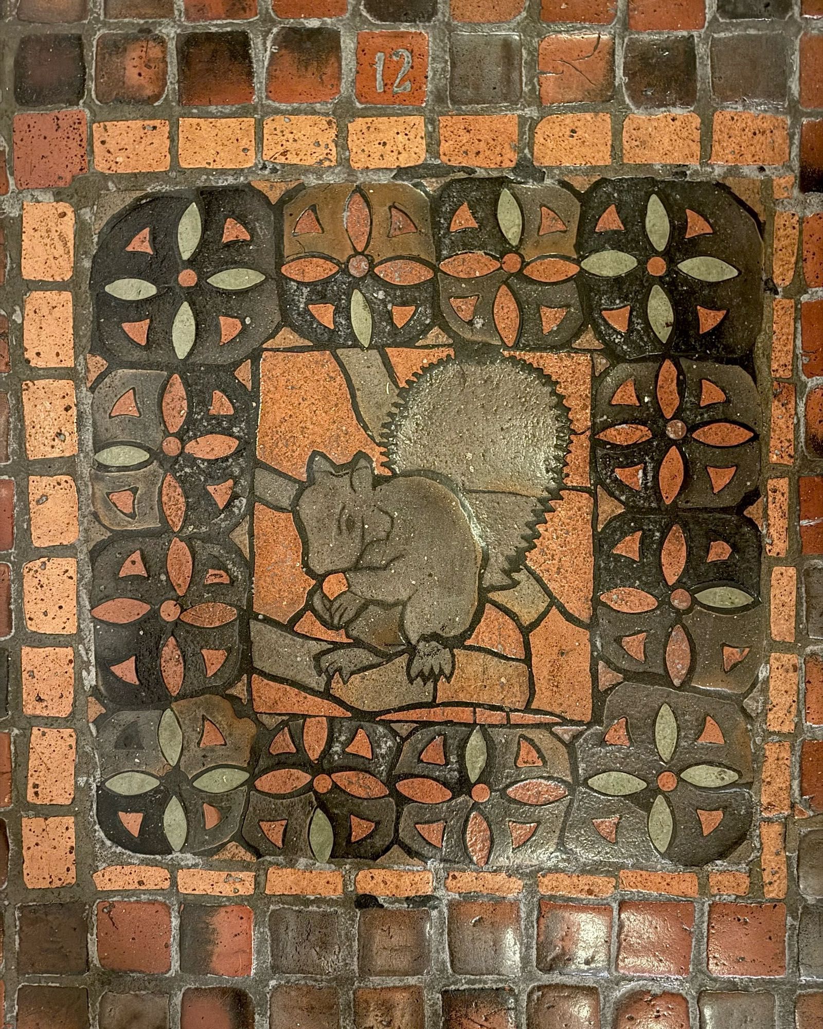 photo of one of the four versions of the grey squirrel floor tile mosaic on the PA Capitol floor, all depicting  squirrel eating a nut on a tree branch: this one numbered “12” and with a decorative border