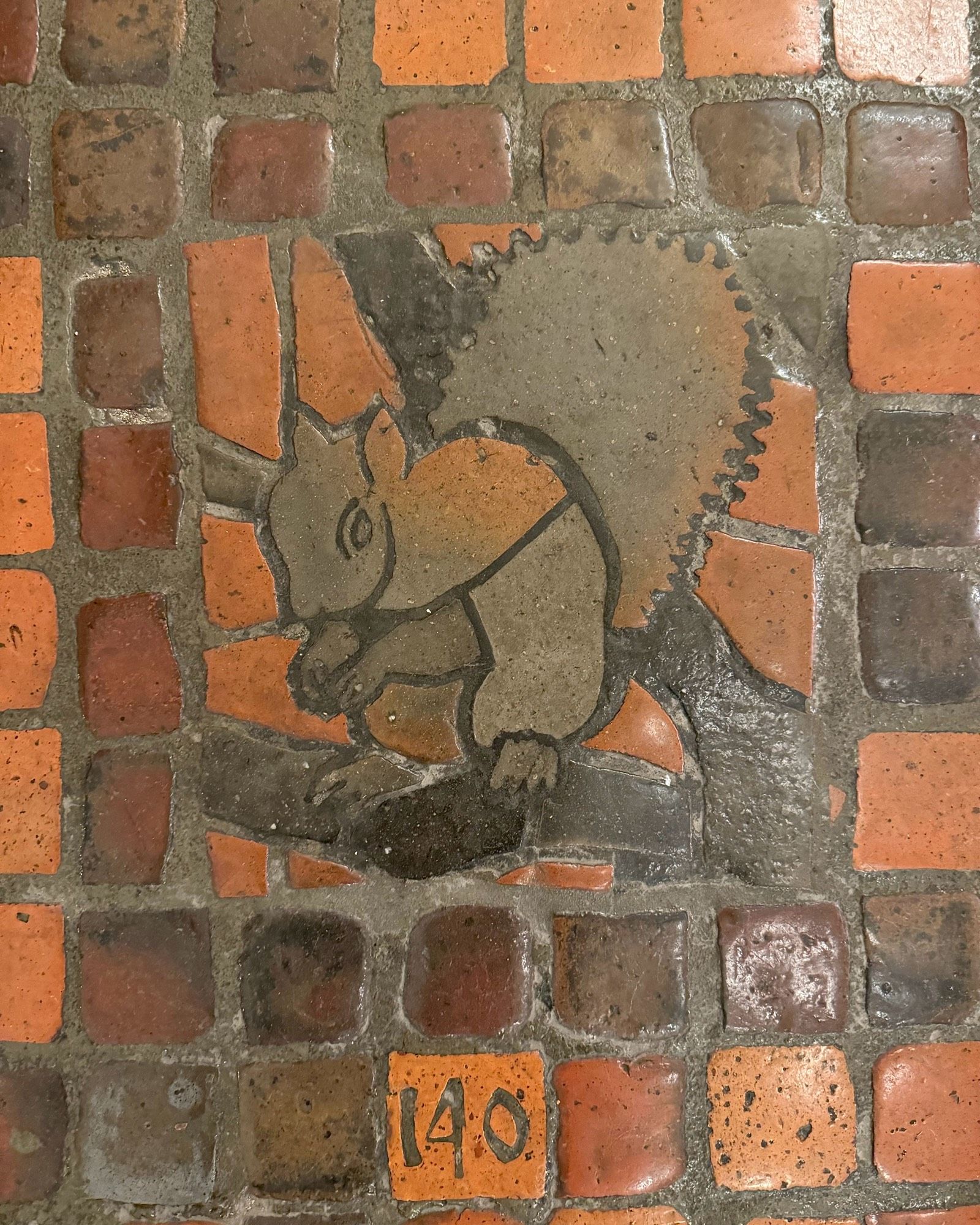 photo of one of the four versions of the grey squirrel floor tile mosaic on the PA Capitol floor, all depicting  squirrel eating a nut on a tree branch: this one numbered “140”