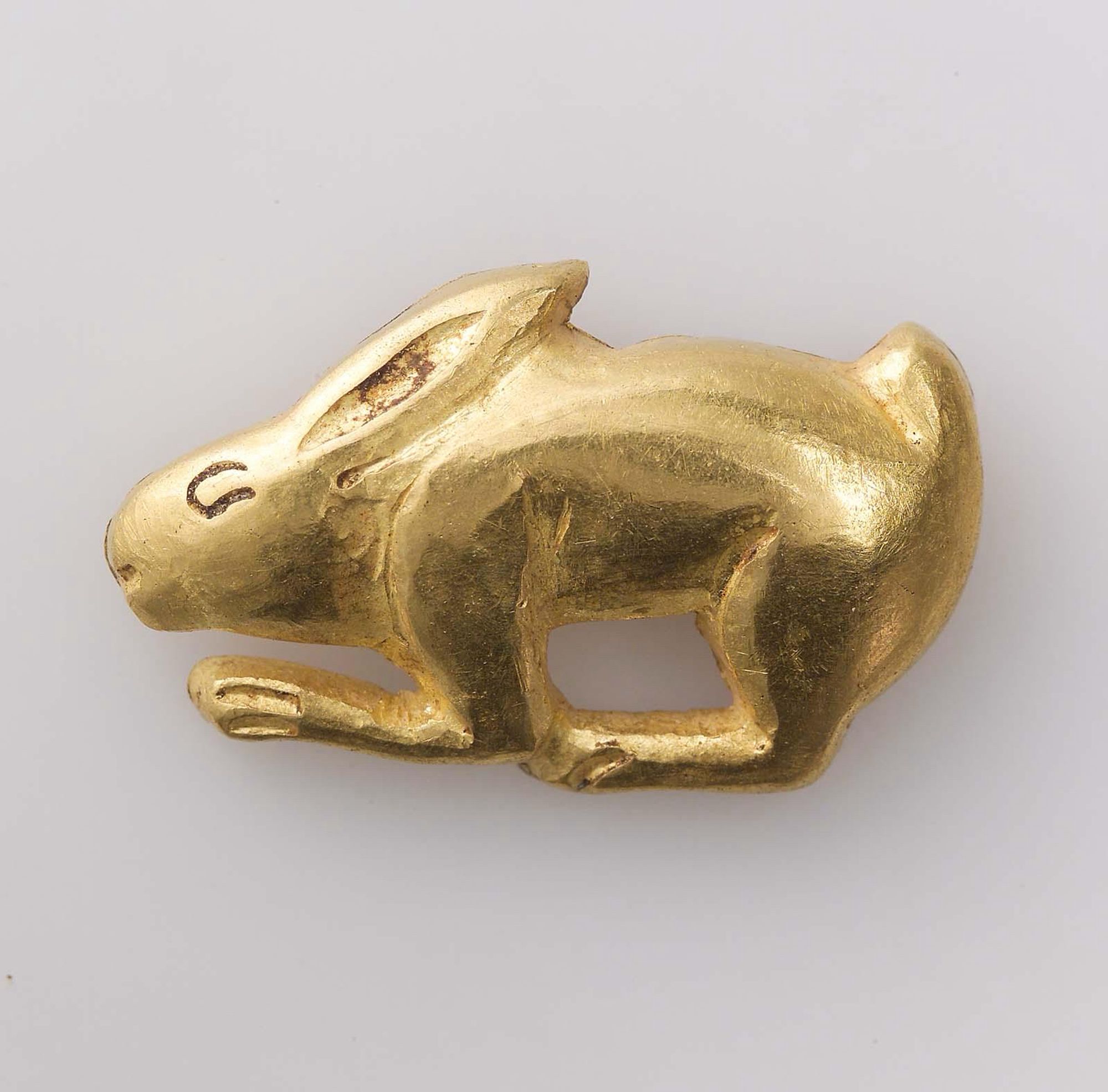 official museum photo of object on white background: small gold ornament in the form of a crouching rabbit in side profile