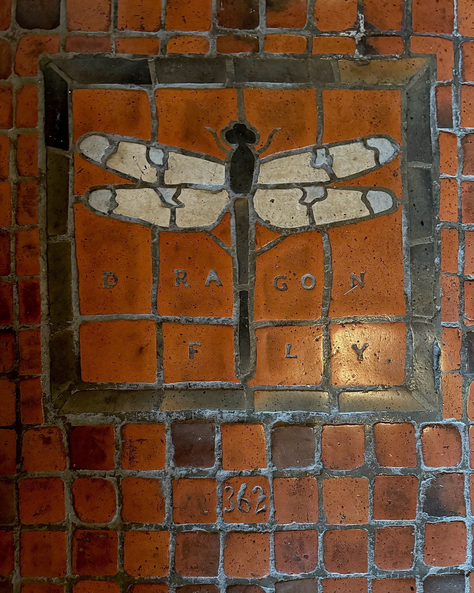 photo of the floor tile mosaic: dragonfly design with black body and white wings with blue spots, square frame, labeled “DRAGON FLY” & numbered “362”