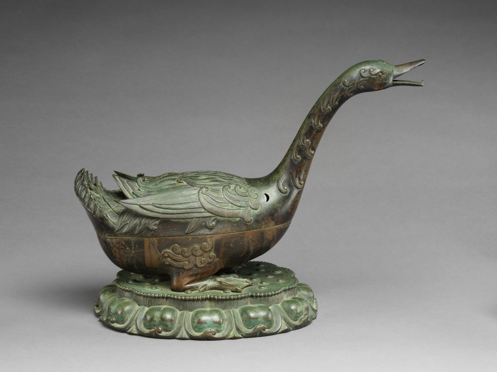 official museum photo of the object on grey background, side profile 1: incense burner in the form of a sitting goose with neck outstretched, bronze with green patina

“This incense burner is an extremely rare example of an imperially-commissioned bronze from the early Ming dynasty. Cleverly designed so that the fragrant incense is exhaled from the bird’s open beak, the censer artfully combines naturalism, ornamental detail, and a sense of history as it references similar incense burners first created during the Han dynasty (206 BC–AD 220) and revived during the Song (960–1279) and Yuan (1271–1368) dynasties. These earlier examples are typically simpler and less animated. The massive size, lively form, and meticulous details of the current work perfectly demonstrate the new taste and high standards of the early Ming period, when the imperial art patronage reached a new peak.”