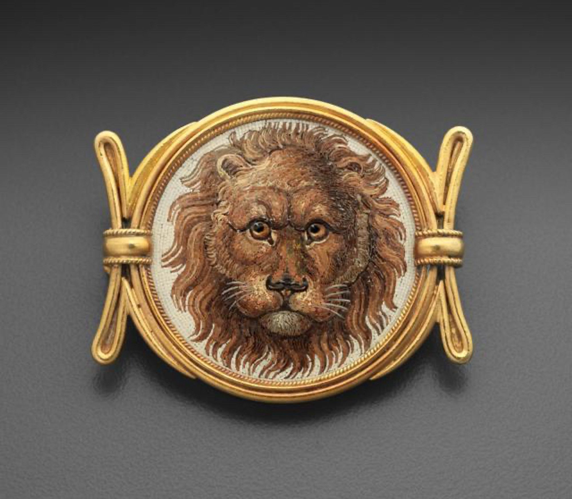 "This brooch features a circular micromosaic panel depicting the head of a lion against a cream ground and set within a gold frame flanked by two tied batons decorated with twisted gold wire. The imagery may have been inspired by the lion head depicted on a Roman floor mosaic discovered in the House of Doves in Pompeii."