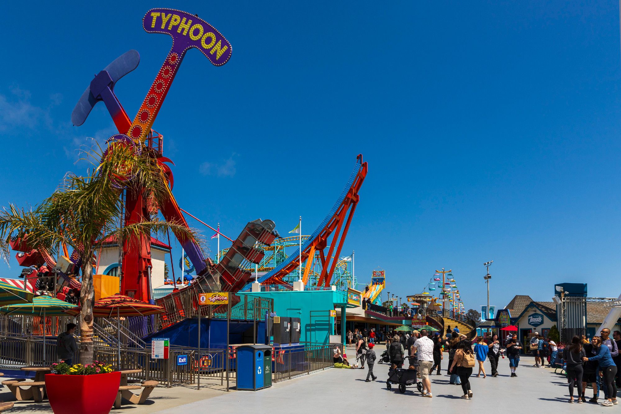 The Santa Cruz Boardwalk under a nearly cloud free, bright afternoon sky. To the left, a ride called Typhoon swings back and forth from left to right. People walk onto the boardwalk/into the park at the right