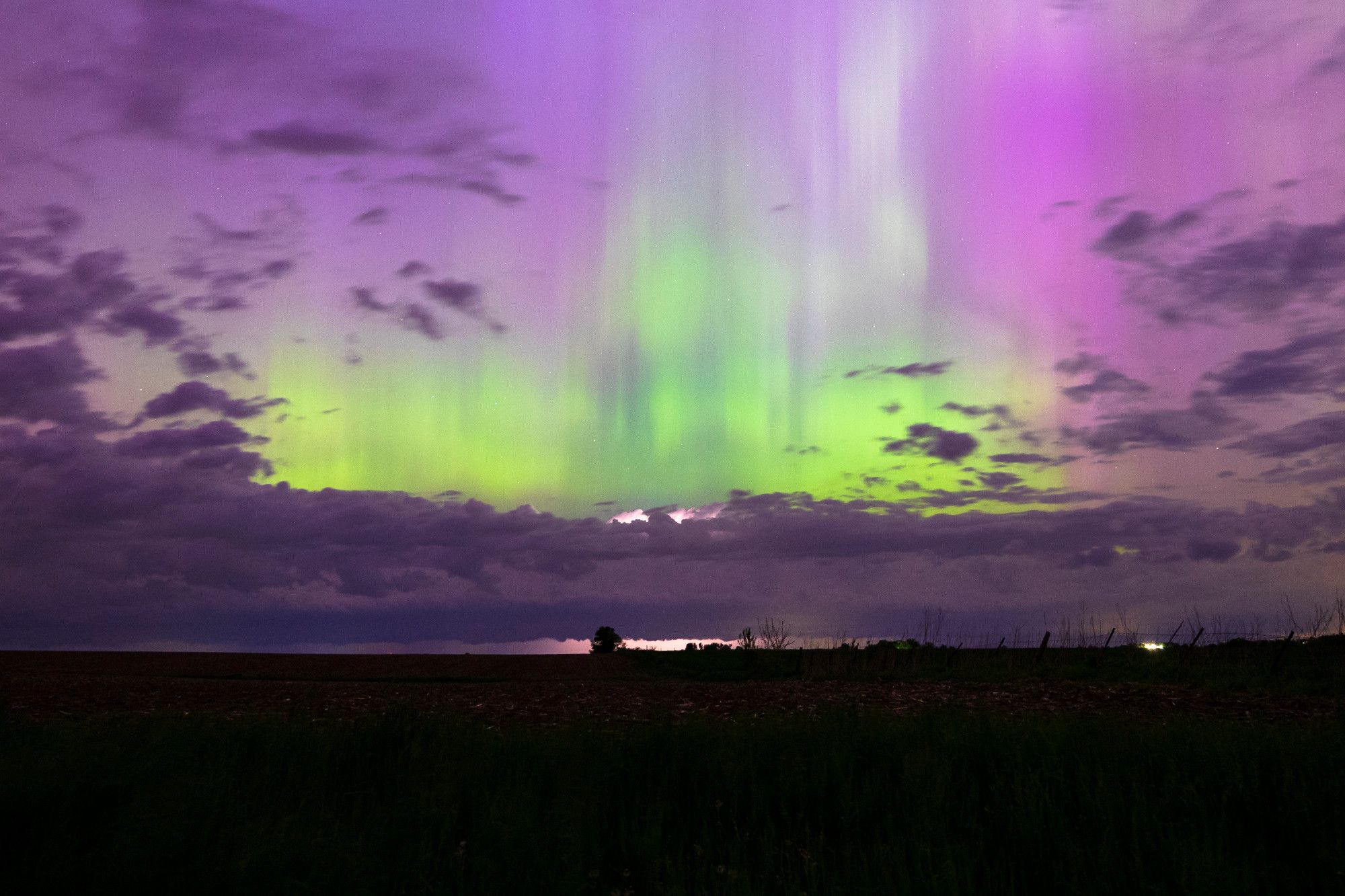 Bright green auroras extending straight upwards above thunderstorm clouds fade into purples the higher you look, dead center of the frame shows a cloud producing lightning above the bottom third of the frame which is darkened by the night