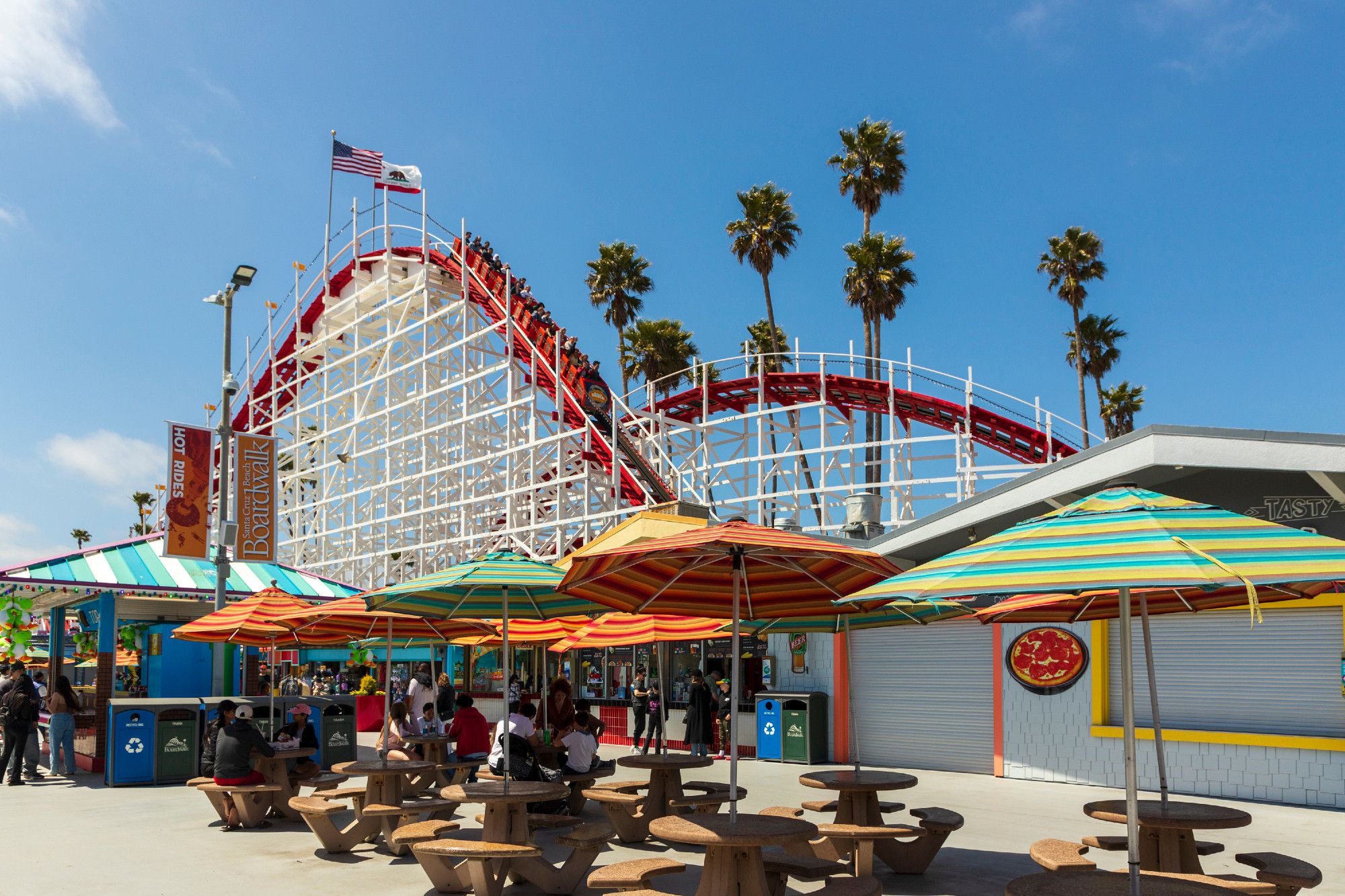 A white and red rollercoaster sees a vehicle head down a first hump, speeding up along the way. Passengers in the rollercoaster are screaming in joy, some with hands held up. Flags of the US and California are steadily blowing in the breeze on top of the ride, palm trees further to the right. In the foreground, umbrella-shaded tables rest unoccupied.