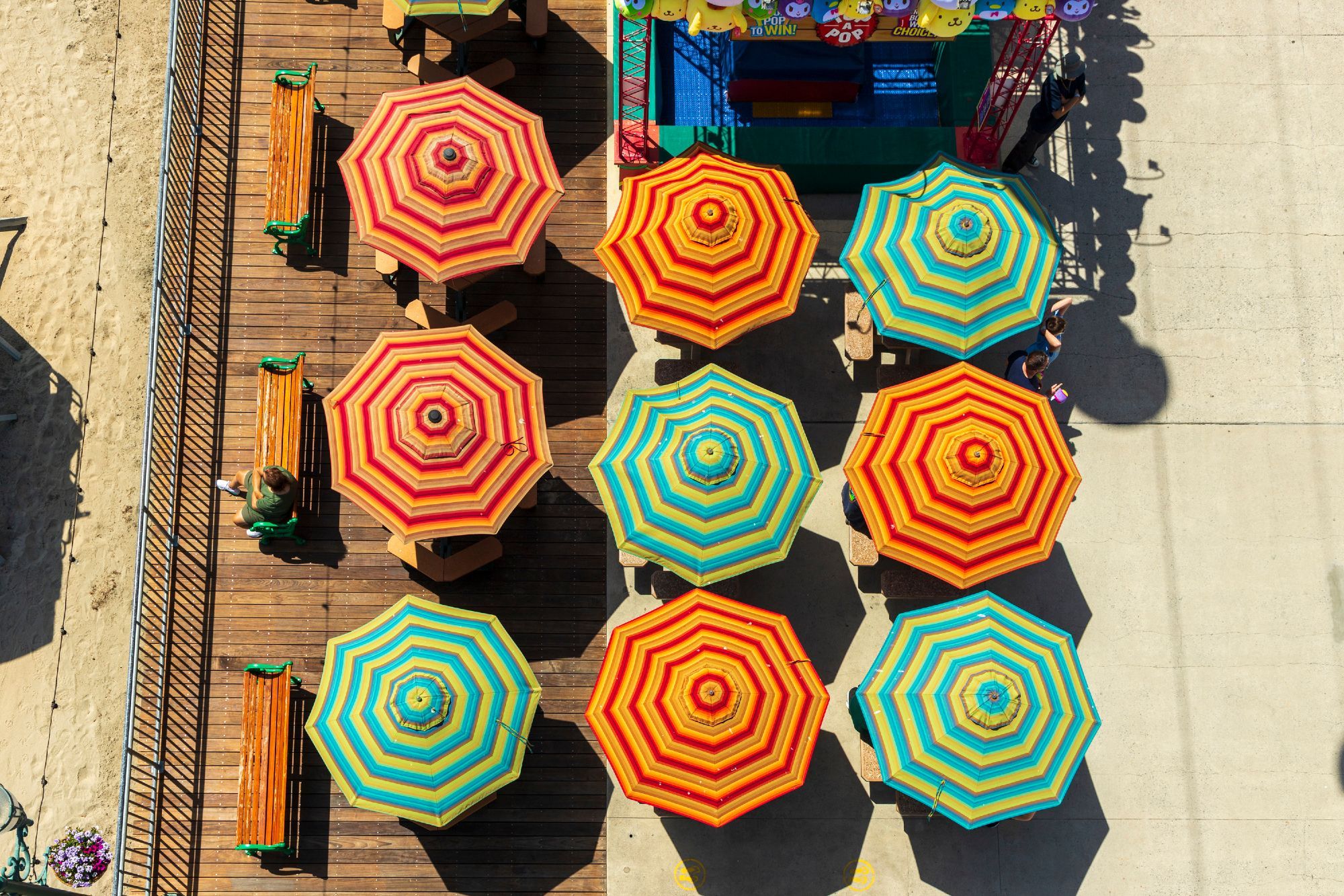 An overhead view of umbrella-shaded tables, taken from a gondola soaring over the boardwalk. The umbrellas are multicolored in ringed patterns of yellows and reds, oranges and reds, sea-greens and reds. Three benches facing the beach are seen to the left of the umbrellas.