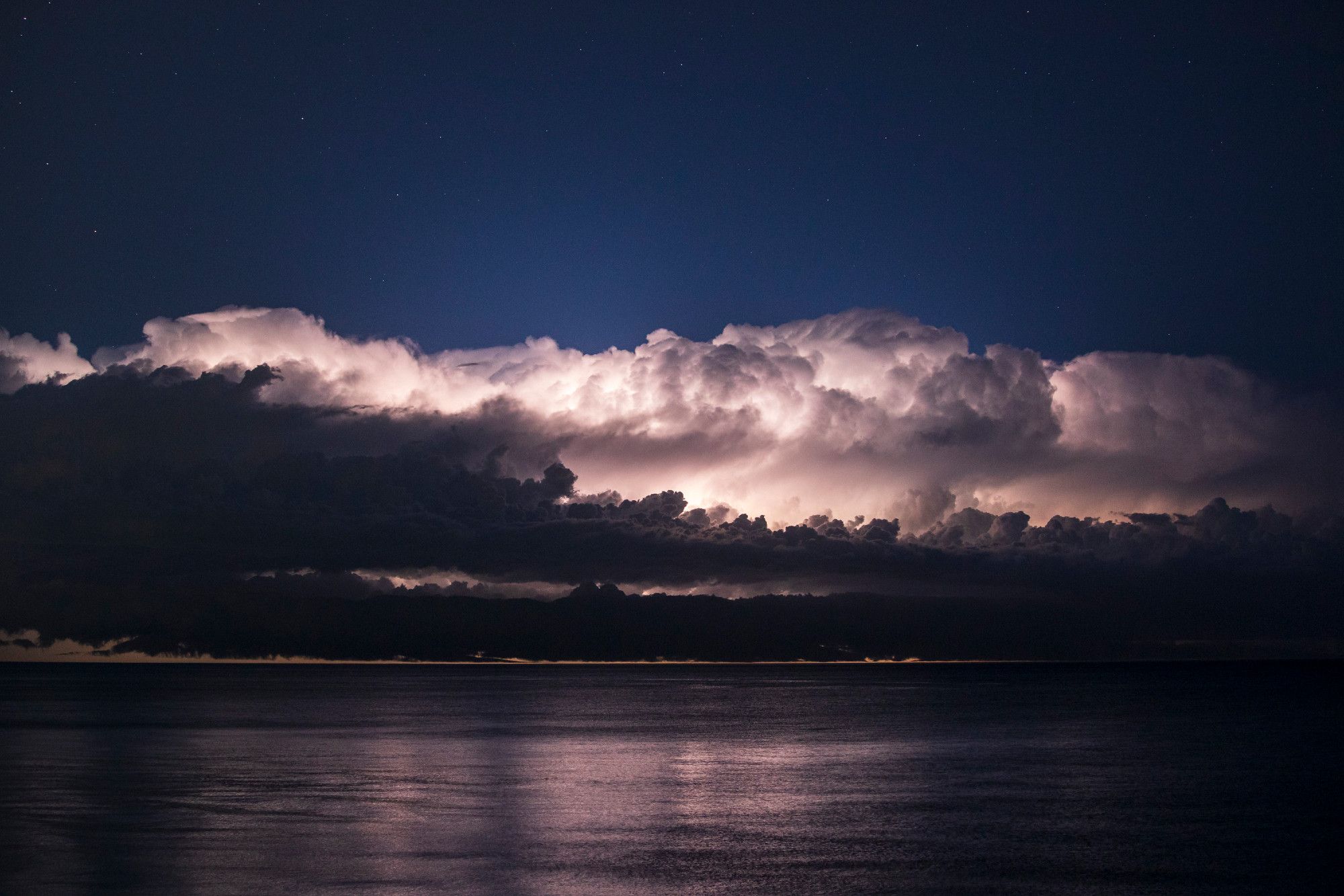 A thunderstorm illuminates the night sky over Lake Michigan. The storm now fills the entire middle third of the frame, with hints of bolts visible between the layers of horizontal clouds in front of the main thunderhead. Said thunderhead is lit up so bright and white it looks like daytime puffy white clouds were dropped into this night scene. The midnight blue sky above has now turned more of a blue hour color from the continuous lightning, and the bottom third lake reflection is much more vivid.