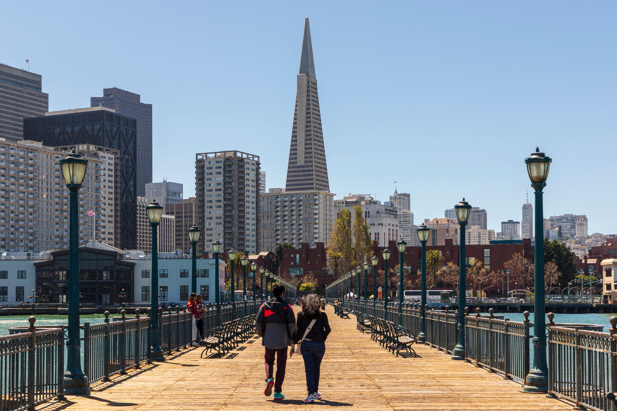 A couple walk hand in hand down a long boardwalk back toward the San Francisco skyline, just about perfectly lined up with the vanishing point that is itself outlined by the boardwalk, streetlights on either side of the boardwalk, and the Transamerica Pyramid building rising from where the vanishing point would be. The two individuals are wearing light jackets, perfect for the weather of the day. There is not a cloud in the sky