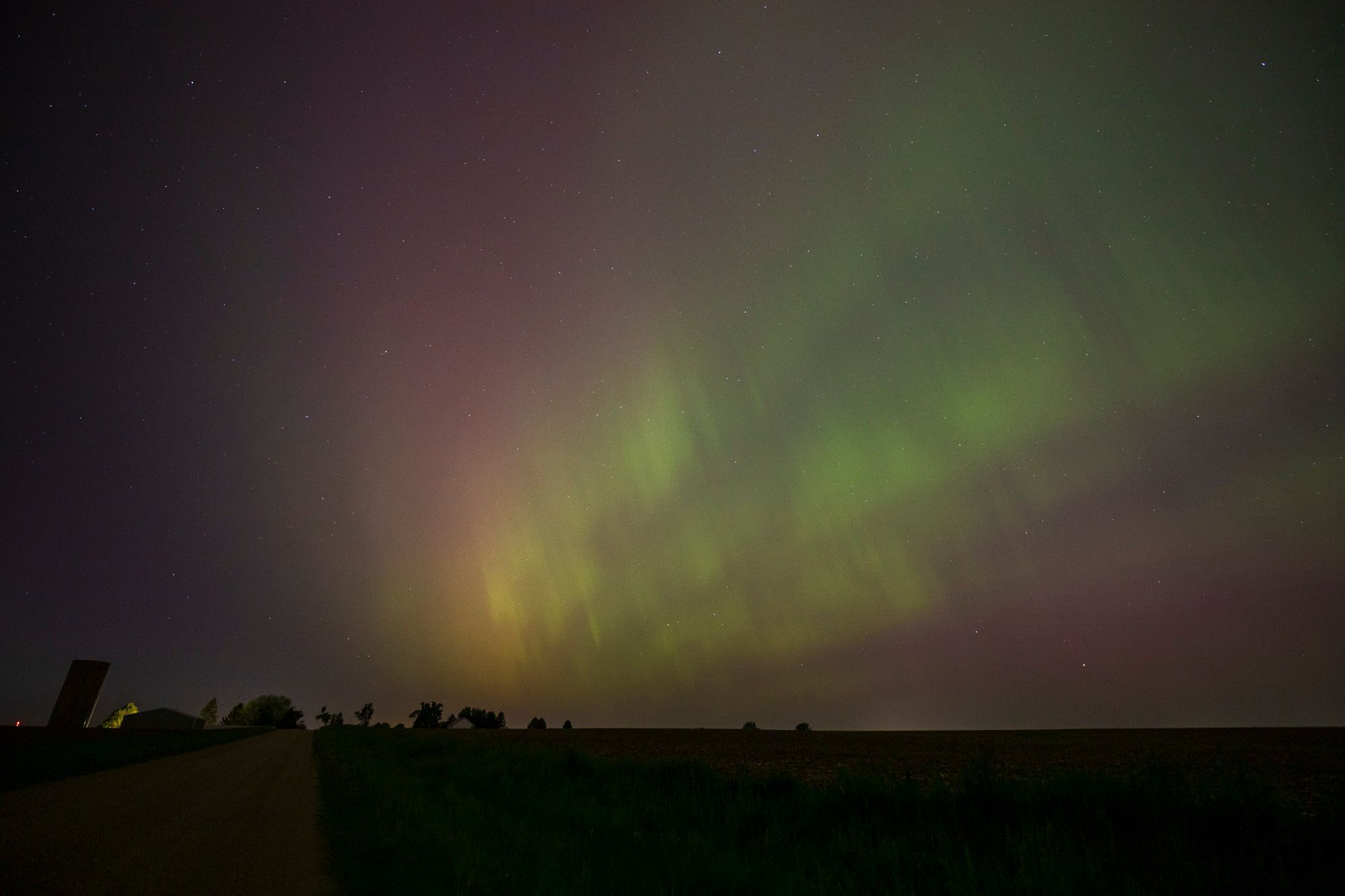 A cloud-free nighttime sky in the countryside, shown off by farm buildings in the distance at the bottom left and a dirt road disappearing into the horizon, are dominated by fingers of bright green aurora extending from center to top-right of the frame