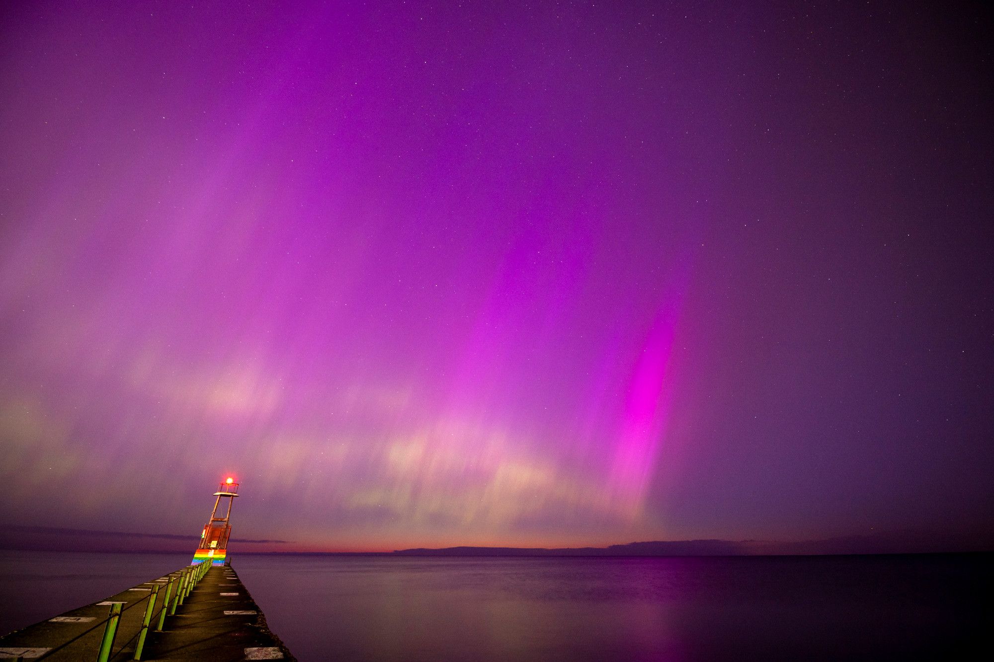 Green waves, bright purple, and darker purple spikes of aurora reflect on a very calm Lake Michigan some time around 4:30 in the morning, with the very very first orange hint of morning light on the horizon just starting to become visible. A short pier on the left extends toward the horizon.