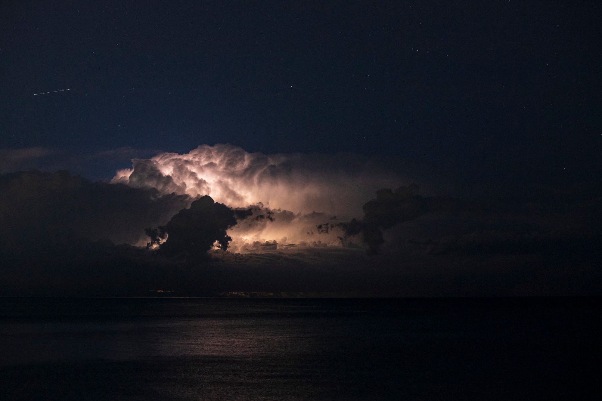 A thunderstorm illuminates the night sky over Lake Michigan. The cloud takes up the middle third of the frame, a bit of white light emanating from a darker, bubbling mass. The top third is a clear midnight blue sky, the bottom third just barely shows a reflection of the light from the cloud