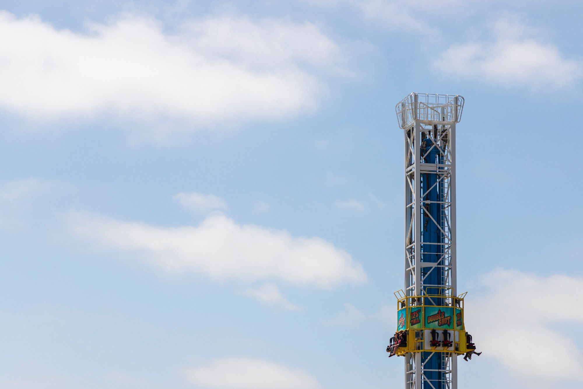 Riders rest at the top of one of those tower falling rides at an amusement part, you know the kind where you go way up and they let you sit there for a minute before dropping you? The drop is about to begin. As this is shot from a gondola the rest of the ride and the ground is not visible, all that is in the background are afternoon blue skies with puffy, white clouds. Are the riders a mile in the sky? Hard to tell.