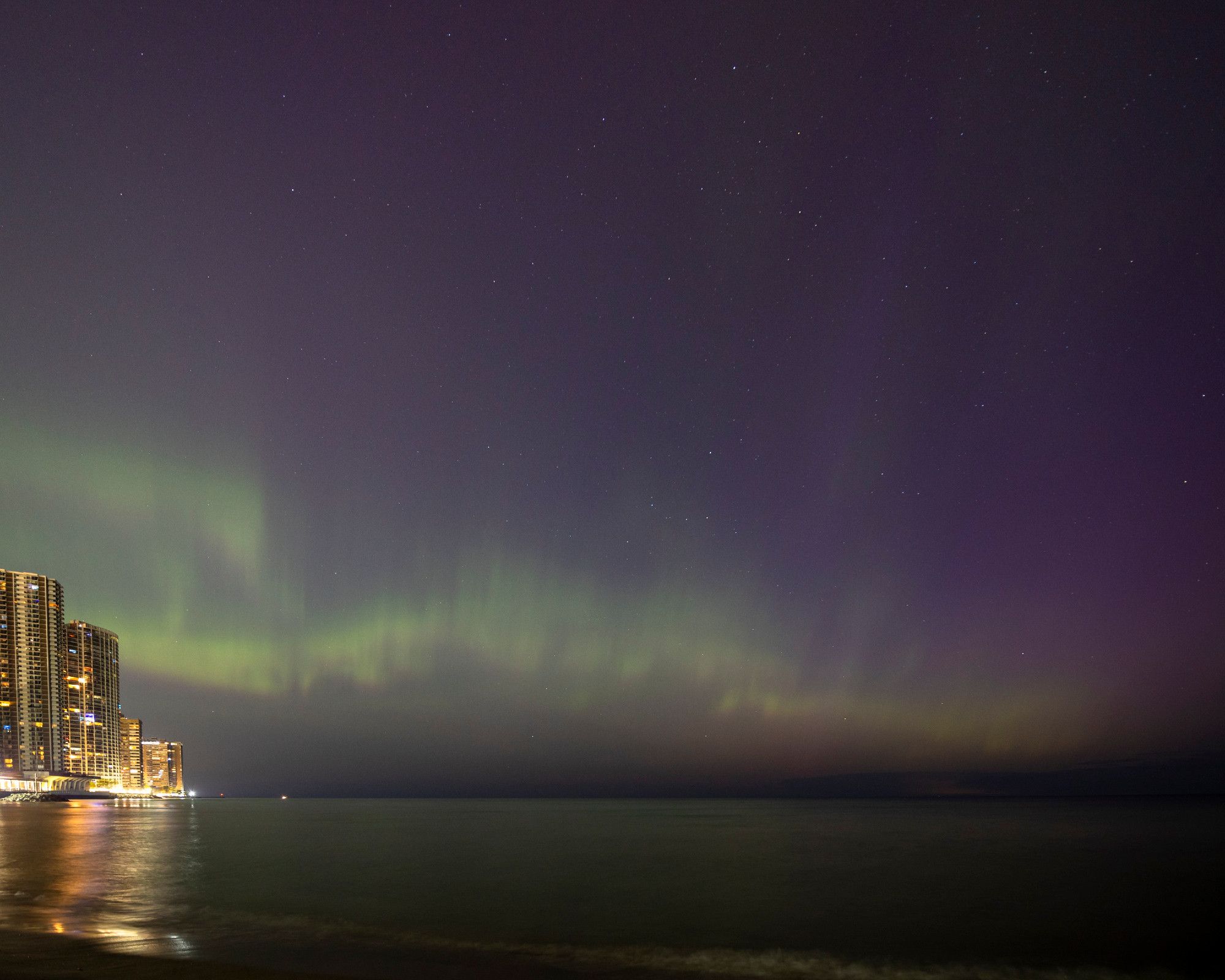 With the skyline of highrises along the Lake Michigan shore of Chicago's Edgewater neighborhood brightly showing at the bottom left, bold streaks of wavy green auroras dance above the horizon over the rest of the frame, gradually fading to dark toward the top