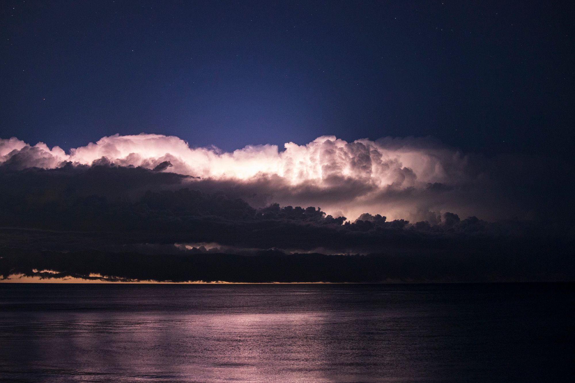 A thunderstorm illuminates the night sky over Lake Michigan. Similar to the last image though between the middle third of clouds and bottom third of lake some clear skies are emerging, themselves colored orange due to how far away the lightning is.