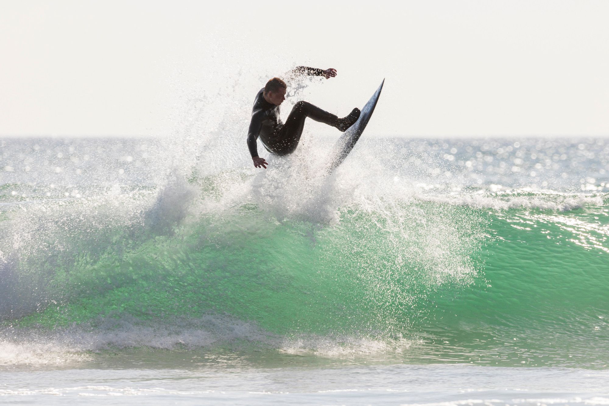 A surfer in a wetsuit successfully tops an ocean wave crashing ashore. The wave is at its peak height when it starts folding over on itself, perhaps 7 or 8 meters high. Bits of the wave are splashing all about the surfer as he slides above the wave.