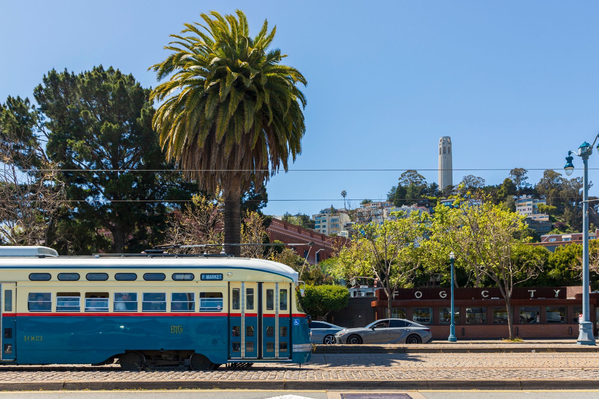 A blue streetcar drives from left to right across the frame along The Embarcadero in San Francisco. A palm tree behind the street is lined up with the front tip of the car, standing dominant above the shorter trees around it. Further to the right and well off in the distance, Coit Tower stands against a cloud-free blue sky