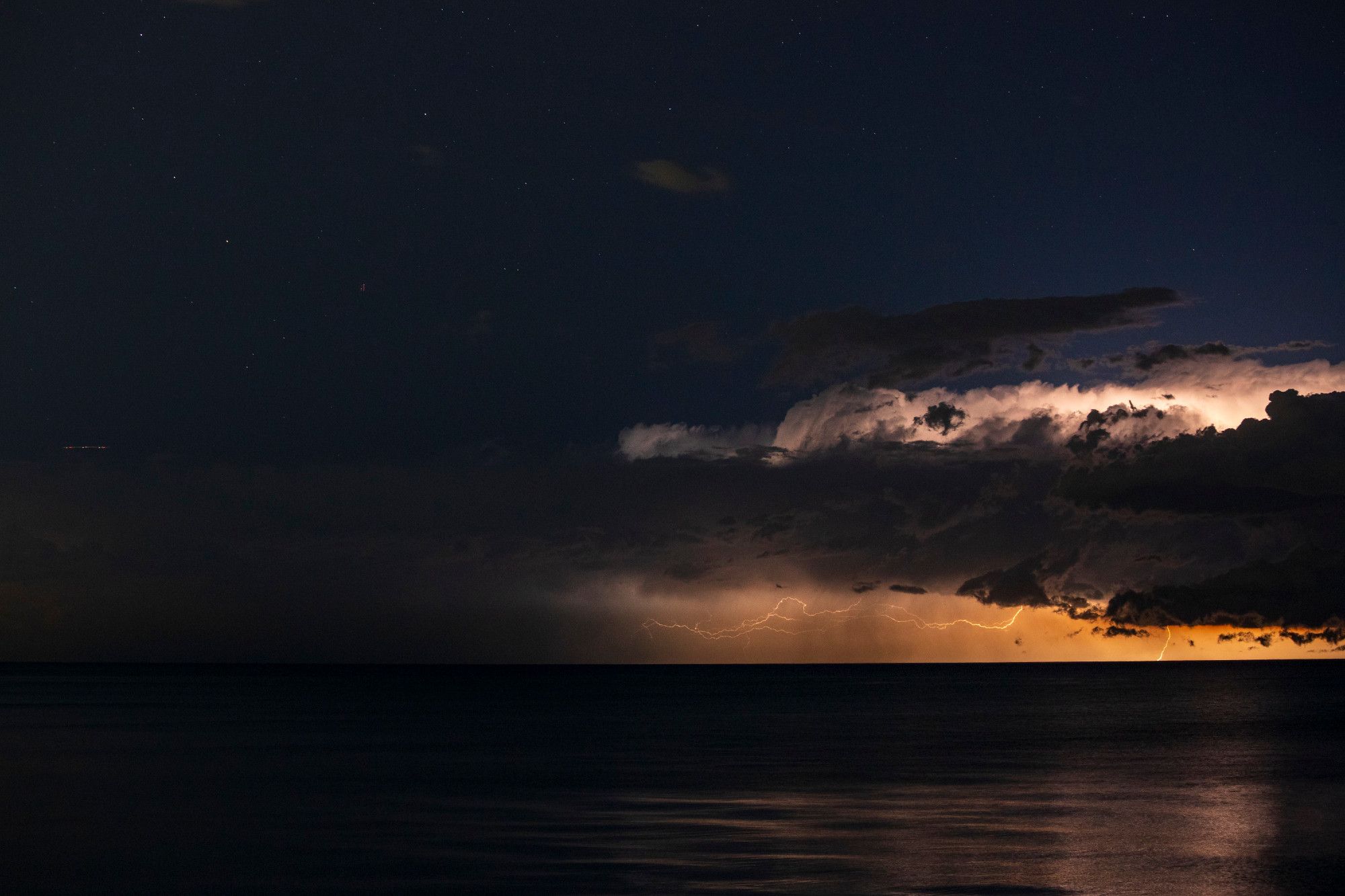 A thunderstorm illuminates the night sky over Lake Michigan. It's departing the frame from left to right, midnight blue and darkness once again taking over the top third and left half of the frame. Bolts of lightning set against an orange sky reach backwards from right to left and toward the darkness. The reflections off the now calming lake appear weaker as the storm comes to an end.