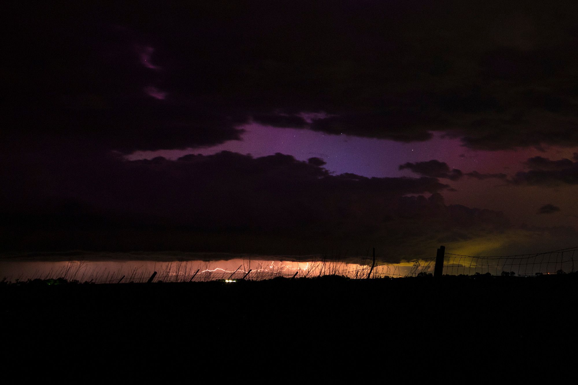 A darkened foreground sits below a yellow-white lightning bolt from the center of the frame jutting to the left, itself below an otherwise mostly cloud-darkened night sky with a narrow slit just above showing off the purple hues of ongoing auroras