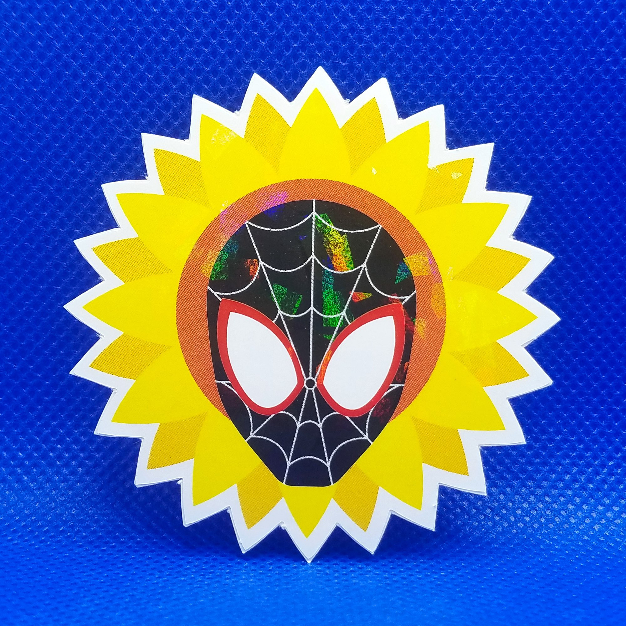 A closeup photo of a holographic sticker depicting the mask of Miles Morales’ Spider-Man surrounding by a sunflower on a blue background. The holographic effect shows prismatic rainbows across the surface of the sticker.