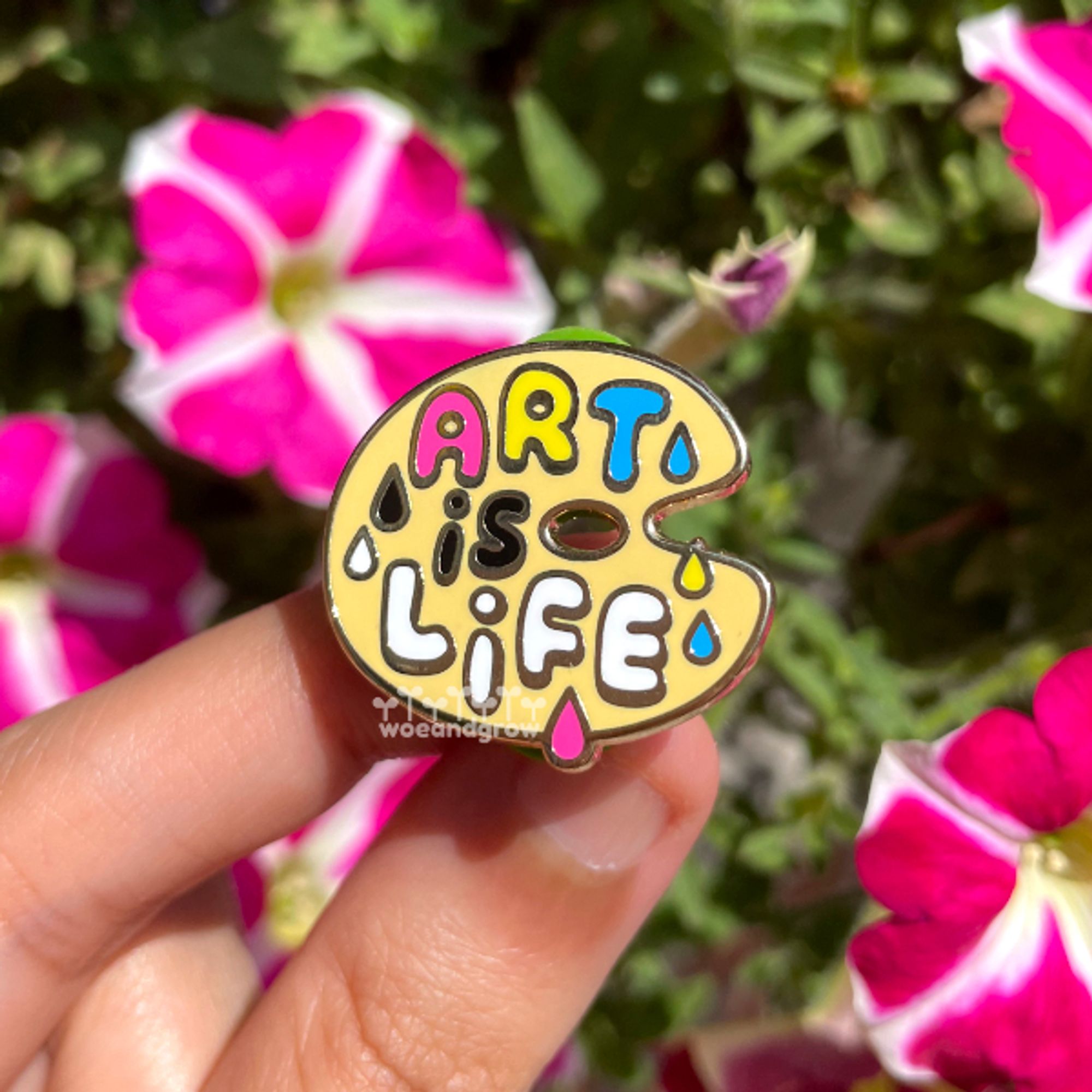 Enamel pin design of an artist pallete with the words "Art is Life" spelled across it as if in paint in CMYK colors. Held in hand over an out of focus background of plants and flowers.