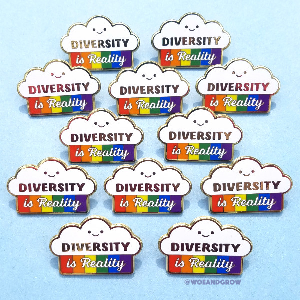 Closeup photo of my Diversity is Reality enamel pins laid out on a light blue background