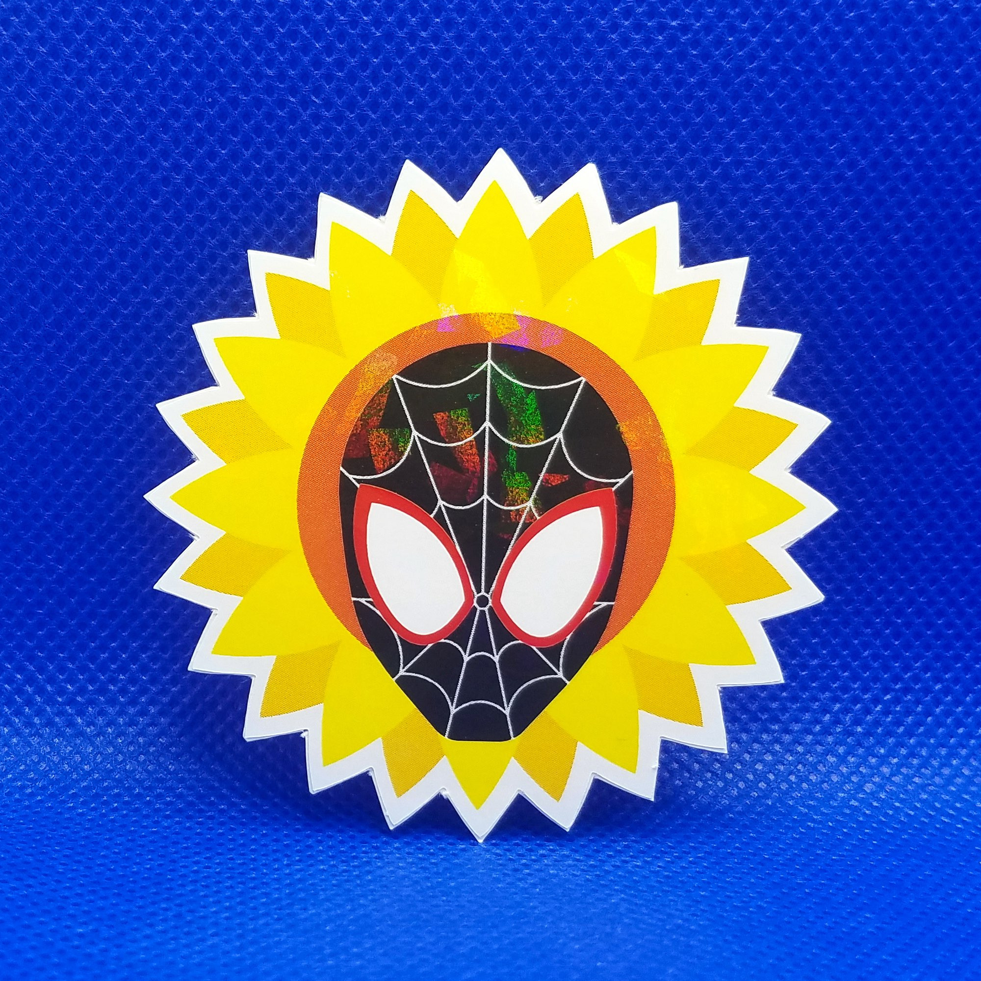 A photo of a holographic sticker depicting the mask of Miles Morales’ Spider-Man surrounding by a sunflower on a blue background. The holographic effect shows prismatic rainbows across the surface of the sticker.