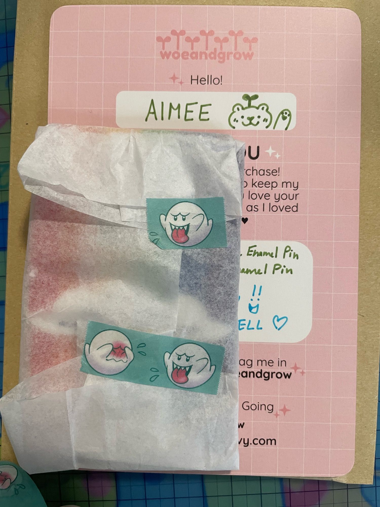 Two enamel pins wrapped in tissue paper and secured with my Blushing Boo washi tape on top of the card insert and packaging on my desk