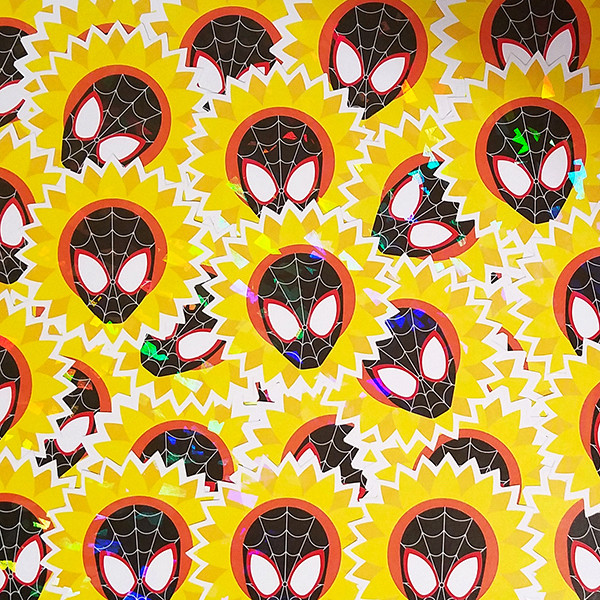 A photo of a pile of holographic stickers depicting the mask of Miles Morales’ Spider-Man surrounding by a sunflower. The holographic effect shows prismatic rainbows across the surface of the stickers.