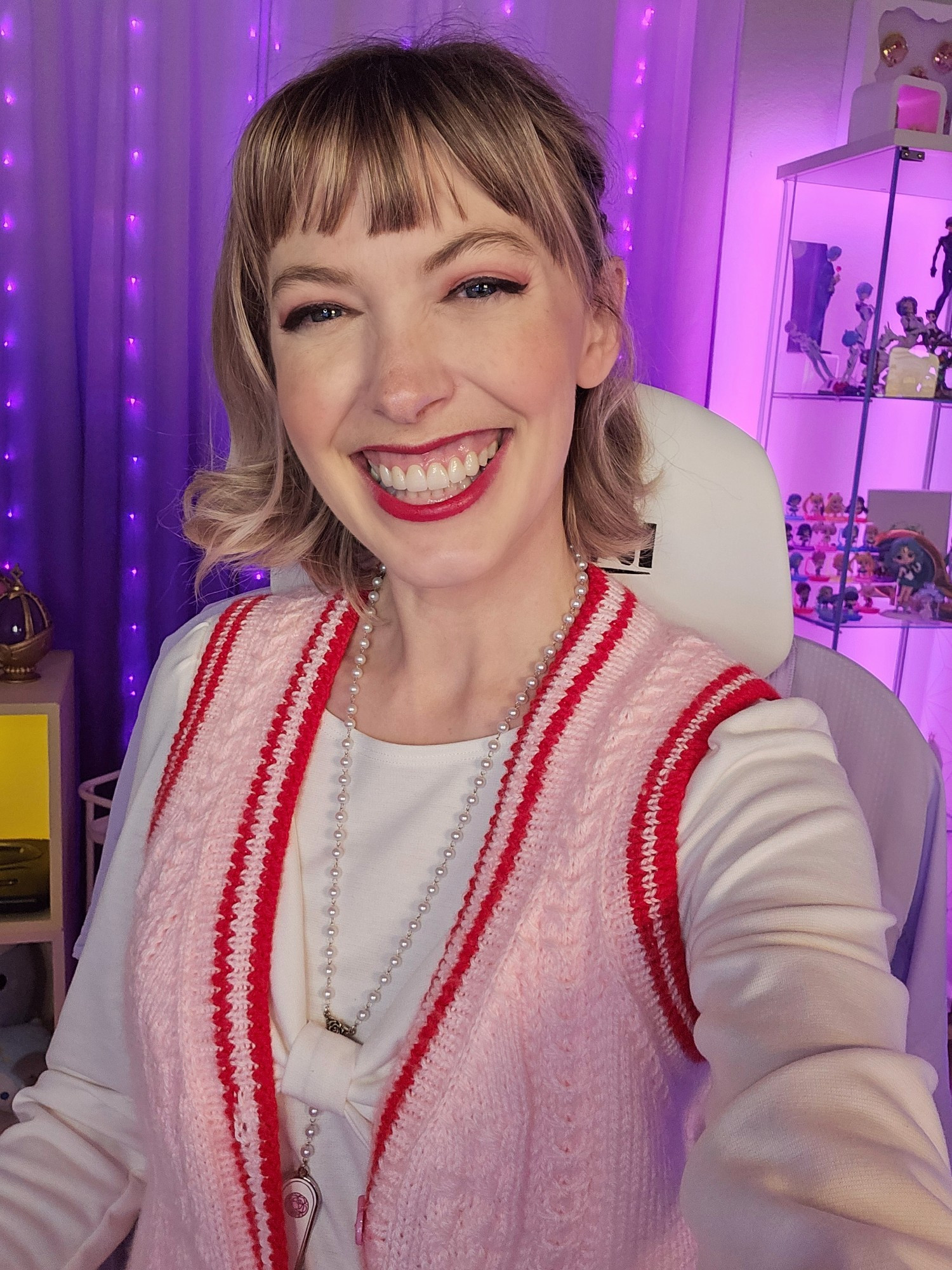 A selfie of me in my streaming room. I'm surrounded by lots of purple lights. I'm wearing a pink sweater and a white dress and a big ol smile!