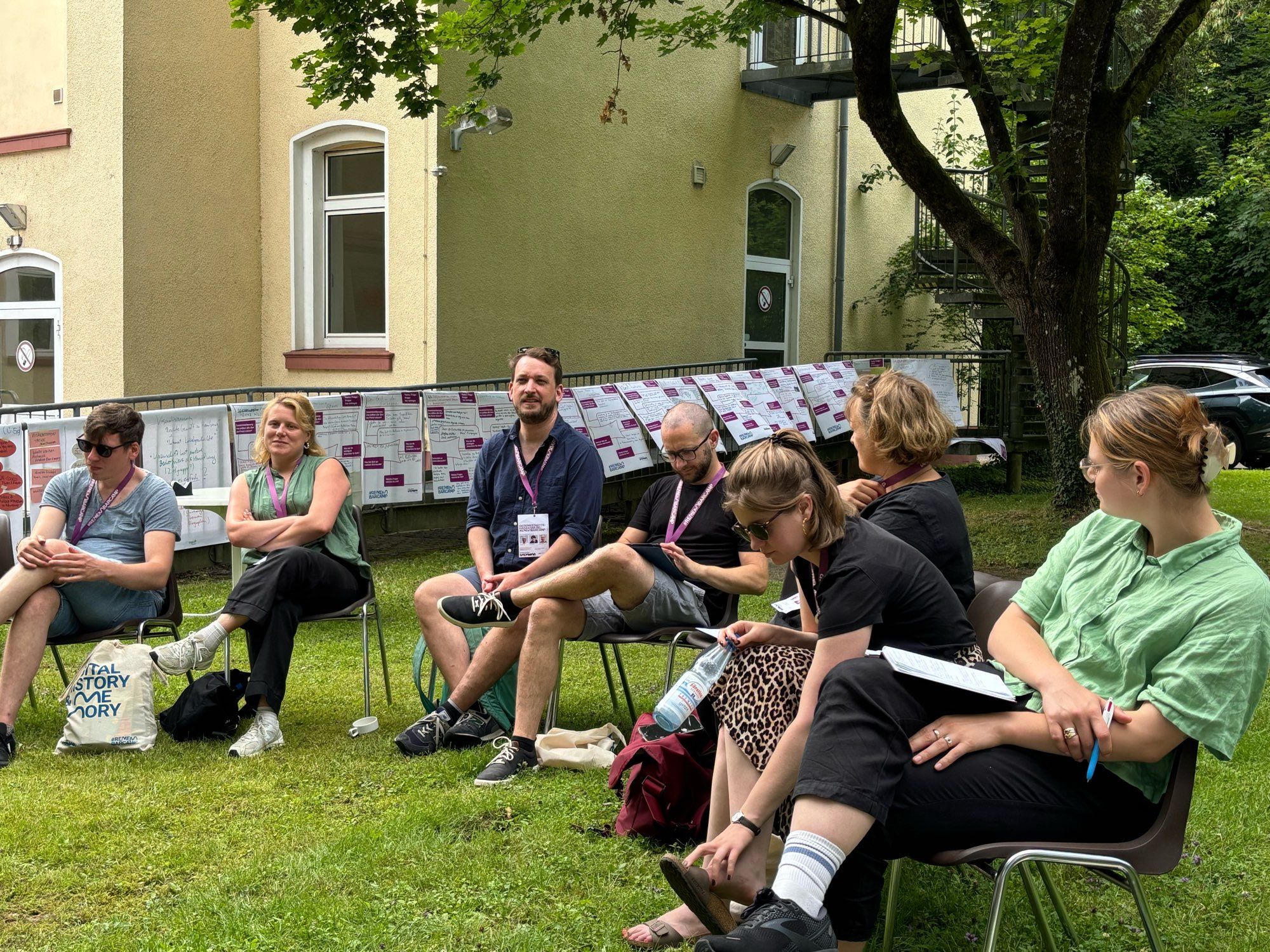 Eine Gruppe von Personen sitzt auf Stühlen auf dem Rasen.  Im Hintergrund eine gelb gestrichene Hauswand und Plakate mit Notizen aus Sessions.