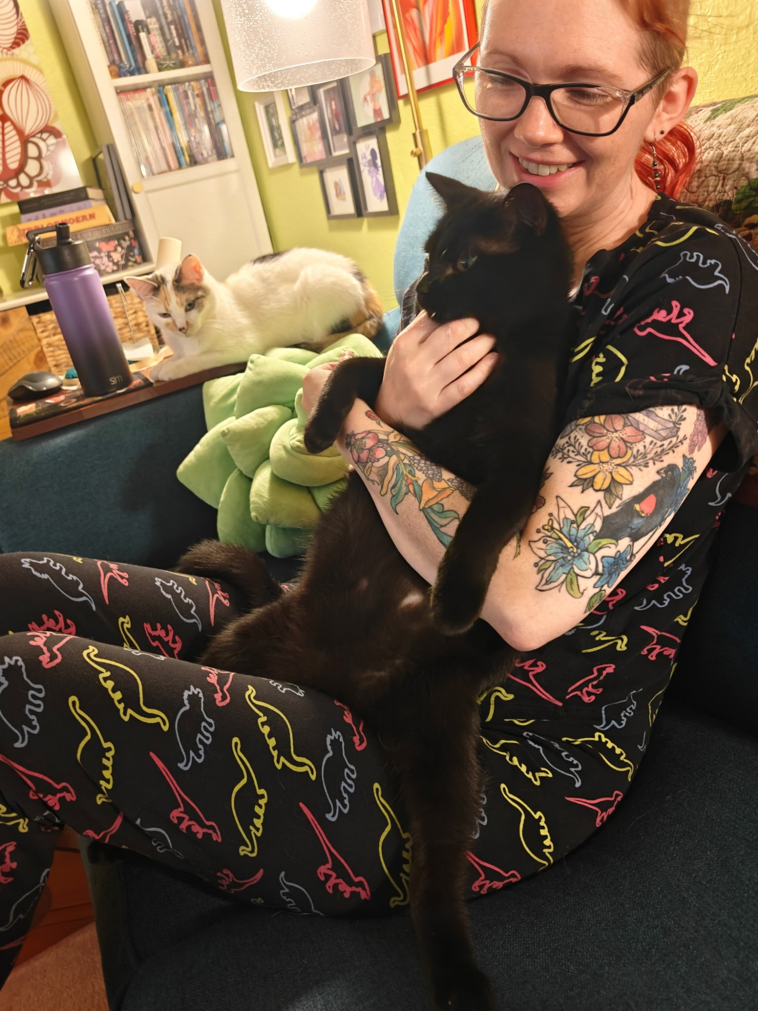 A pale white woman with red hair in dinosaur print pajamas sits on a couch with a black cat in her lap. The cat is straight up sitting like a toddler. He has one back leg on either side of her thigh, and is leaning his back against her chest. She has one arm wrapped loosely around his midsection, over which his front paws drape casually toward the floor.