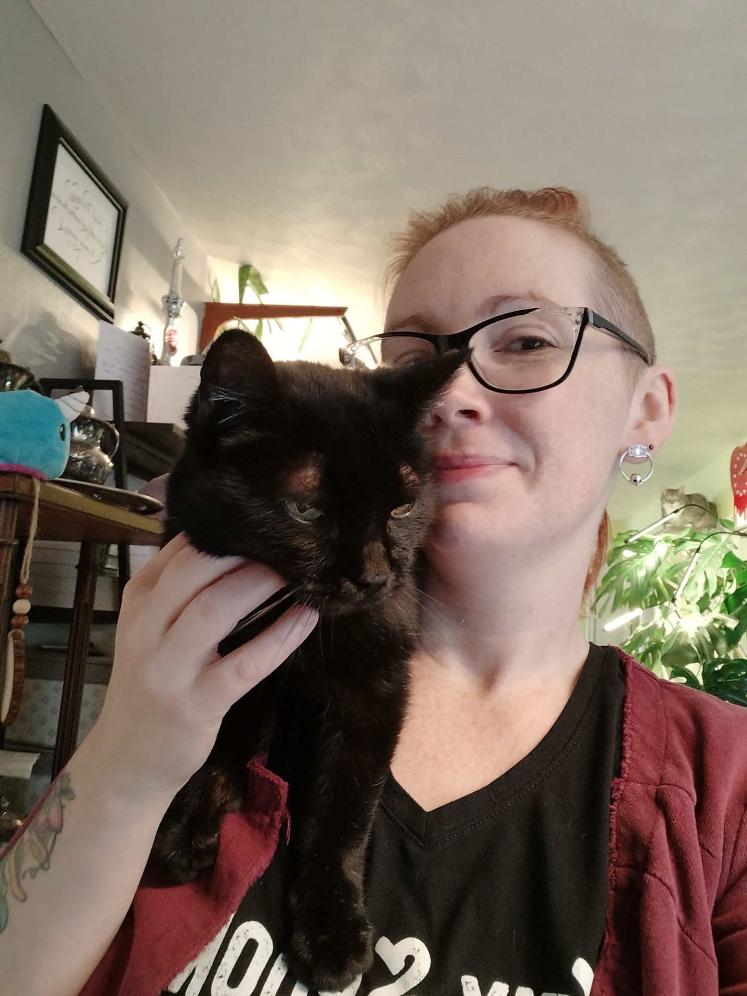 A pale white woman with red hair looking resignedly at the camera while petting the small black cat perched on her shoulder.