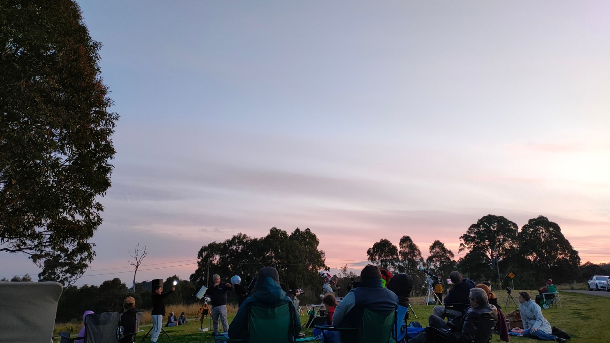 A group of people sitting on a hill in a park at sunset. Everyone is rugged up and has telescopes ready for nightfall.