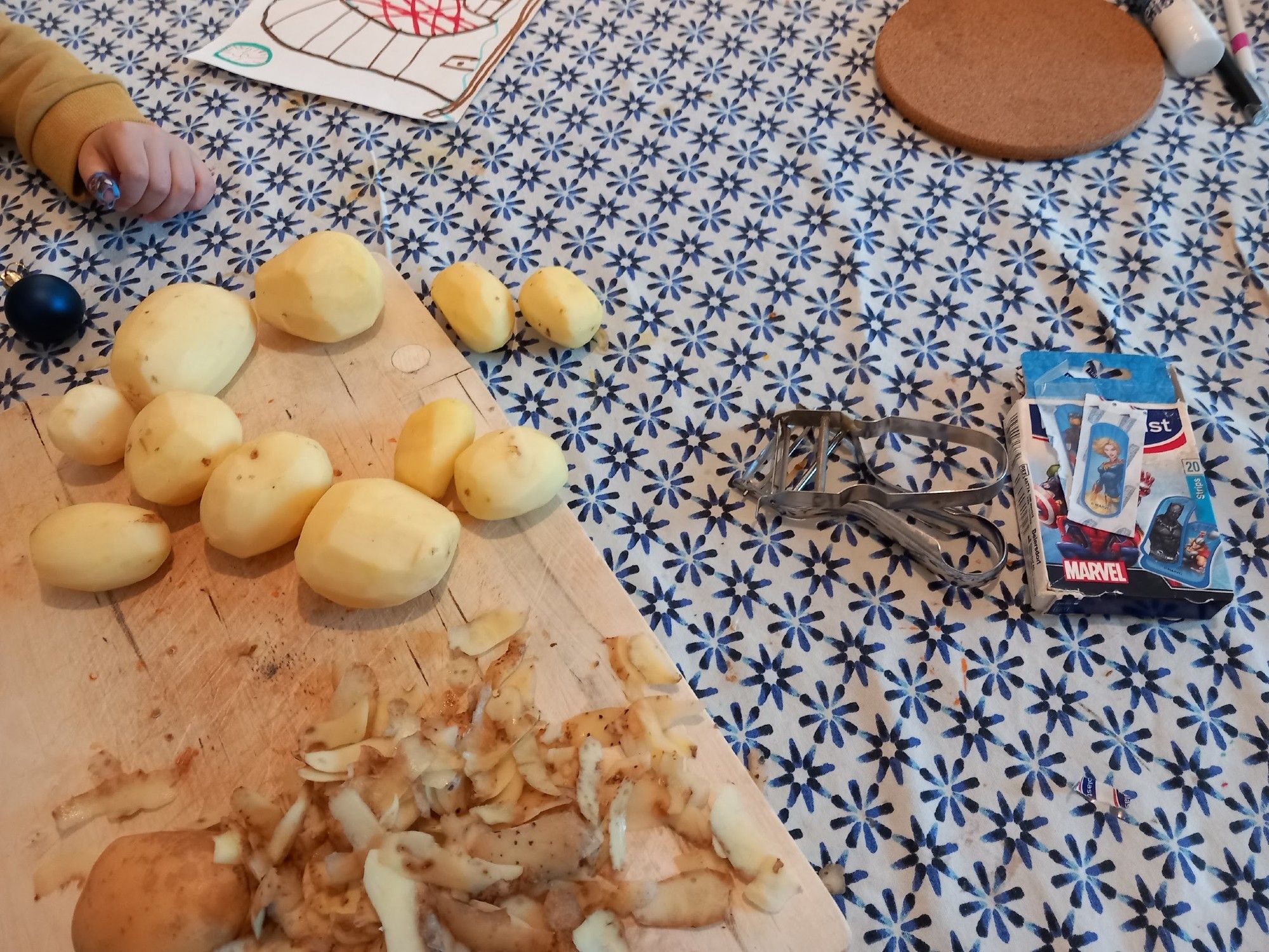 Photo d'une table avec une nappe. En haut a droite une main d'enfant avec un sparadrap sur le doigt. Une planche avec des patates pelées. Deux couteaux économes. Un paquet de sparadrap "super héros".