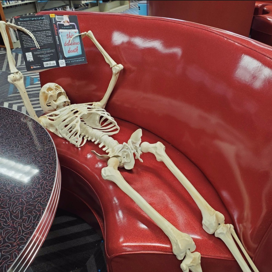 A skeleton leaning on a bright red bench straight out of a 1950s diner or ice cream shoppe. The skeleton is reading a book about death.