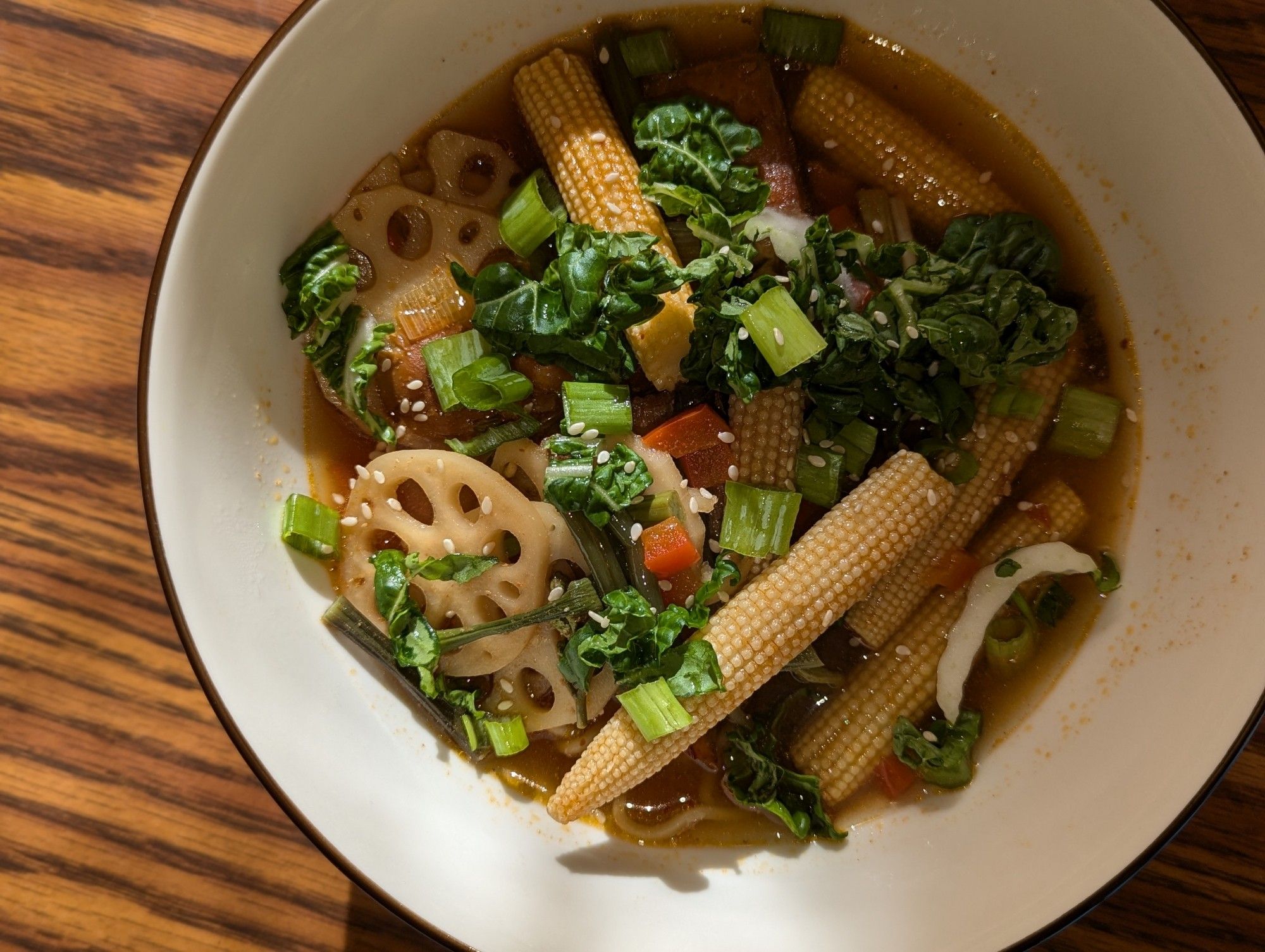 Ramen, in a large white bowl. Baby corn, bok choy, lotus root, red pepper, fresh green onion and sesame seed sit within a rich brown spicy miso broth. One lone noodle pokes up to the surface. The bowl is off center enough that you know the photographer is a novice.