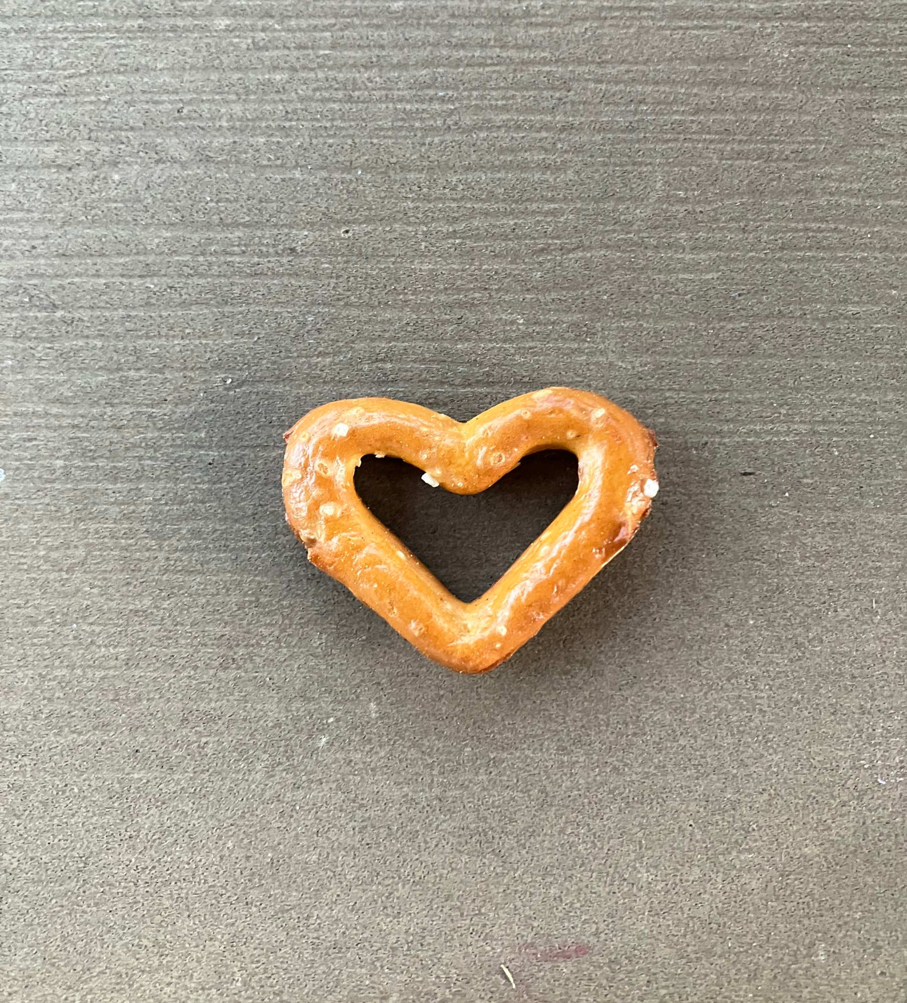 Auf dem Bild ist ein kleines Salzbrezelstück zu sehen, das in der Form eines Herzens gebacken ist. Es liegt auf einer ebenen, graubraunen Holzoberfläche. Die Brezel hat eine goldbraune Farbe mit vereinzelten Salzkörnchen darauf. Das Herz ist gleichmäßig geformt und hebt sich durch seine runde, glänzende Oberfläche deutlich vom Hintergrund ab.

Bildbeschreibung durch KI.