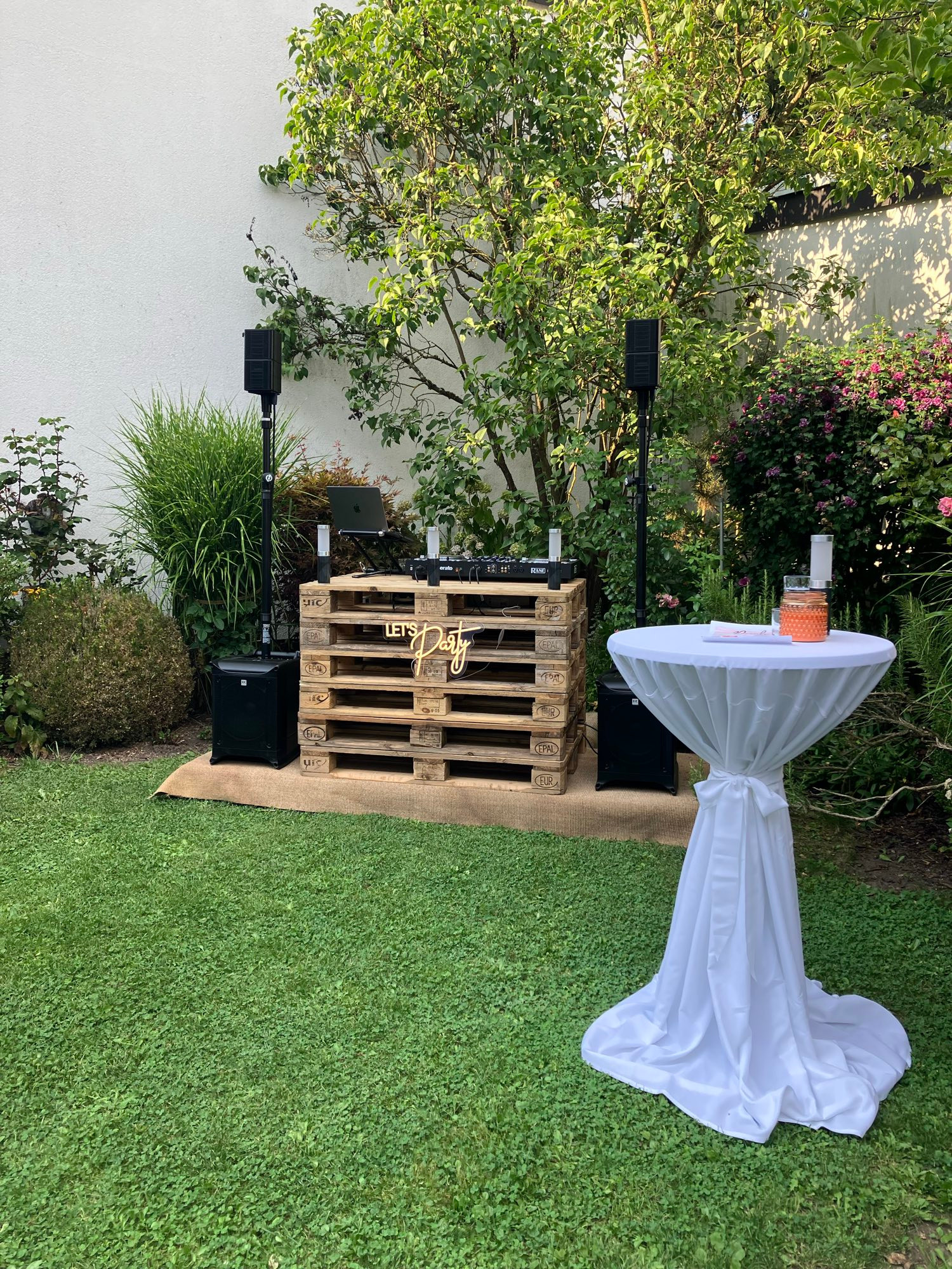 Auf dem Bild ist eine kleine Outdoor-Party- oder Event-Szene zu sehen.
In einem gepflegten Garten mit grünem Rasen steht im Hintergrund ein improvisierter DJ-Tisch, der aus mehreren gestapelten Holzpaletten besteht. Darauf befinden sich DJ-Equipment, ein Laptop und zwei kleine LED-Lichtsäulen. Vor den Paletten hängt ein dekoratives Schild mit der Aufschrift „Let’s Party“ in gelber Neon-Optik. Links und rechts neben dem Tisch stehen auf Stativen zwei Lautsprecher, am Boden zwei Subwoofer.

Rechts im Vordergrund steht ein hoher Stehtisch, der mit einer weißen, bodenlangen Tischhusse bezogen ist. Darauf befinden sich einige Gläser, ein orangefarbenes Windlicht und eine weiße Kerze. Im Hintergrund wachsen Büsche, Sträucher und ein Baum, die eine gemütliche und private Atmosphäre schaffen.

Bildbeschreibung durch KI.