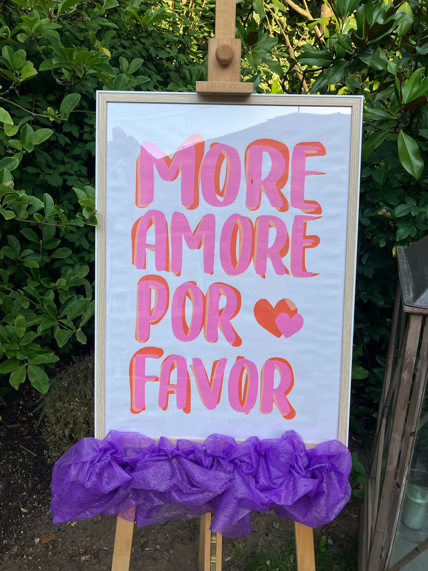 Staffelei mit Bild und der Aufschrift More Amore por favor in einem Garten.