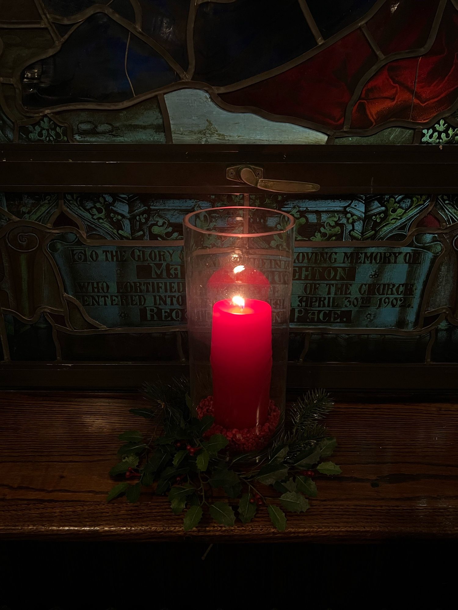 Large red candle quietly burning in a glass case, with a wreath of holly decorating its base.